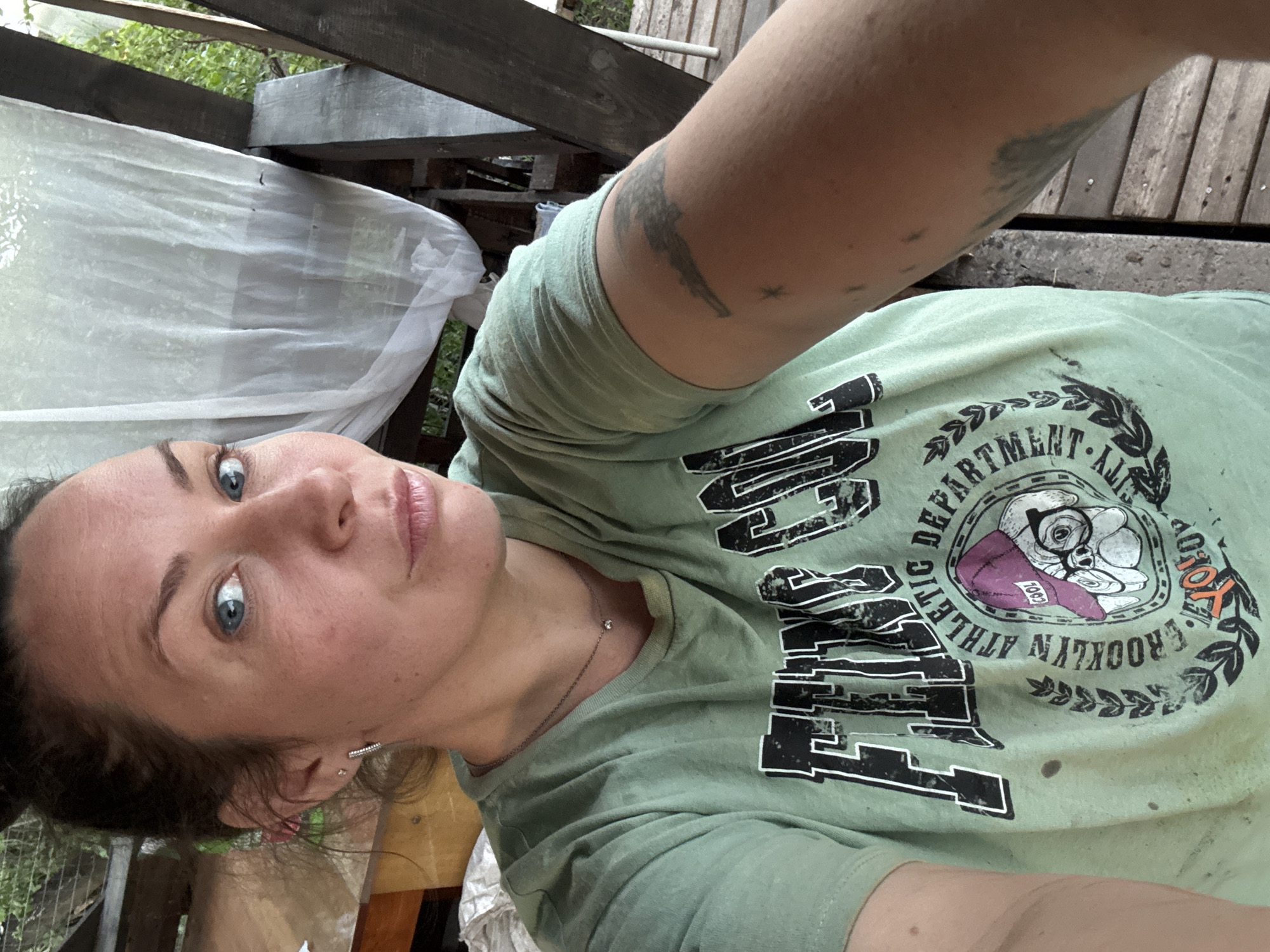 A selfie taken from a low angle featuring a Caucasian woman with light skin and blue eyes. She has dark hair pulled back, exposing a small earring. She wears a light green t-shirt with a black and white graphic print, including the word "Brooklyn" and "Athletic Department," along with what appears to be a stylized dog wearing a hat and glasses. There is a tattoo on her left arm. The background shows a wooden structure or deck, and foliage is visible. A white, semi-transparent material drapes in the top left corner of the image. The setting appears to be outdoors, likely in a backyard or similar space. The location in the city of Chaikovsky, Russia is not determinable from the image itself.