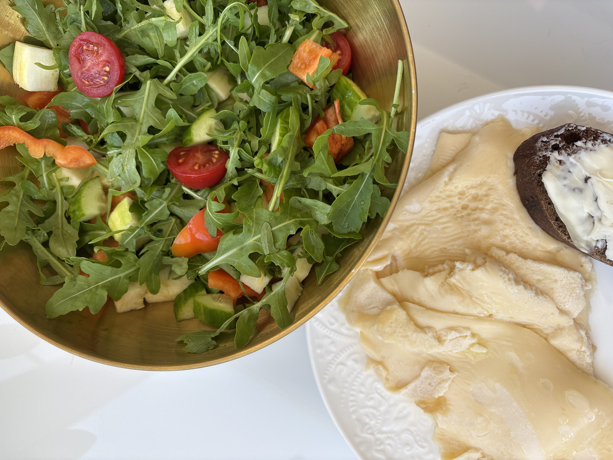 A close-up, overhead shot showcases a meal on a white surface. In the upper right, a golden bowl overflows with a vibrant salad composed of arugula, cherry tomatoes, cucumber, and orange bell peppers. To the left, a white, patterned plate holds thinly sliced, pale yellow cheese, with a piece of dark bread topped with a white spread. The lighting is bright, suggesting an indoor setting. There is no indication of the city of Razdolnoye, Russia, or any people present.