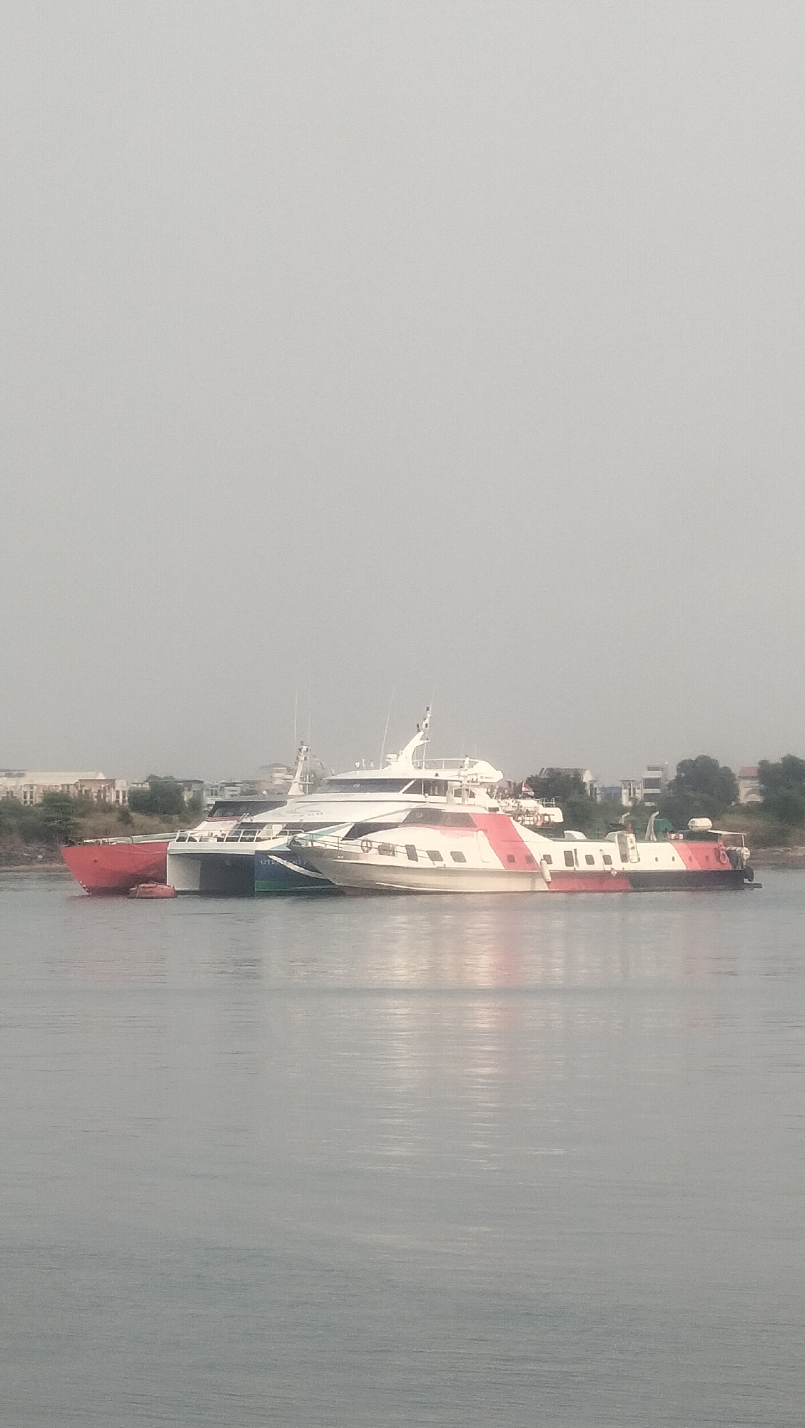 The image shows two catamarans docked side-by-side in the waters of Batam, Indonesia. The catamaran on the left has a red hull and appears to be a passenger ferry. The catamaran on the right is white and red with a more modern design, also likely a passenger ferry. Both vessels are moored close to the shore, where a line of buildings and trees can be seen in the background, suggesting an urban or coastal environment. The sky is overcast, casting a diffused light over the scene. The water is calm with reflections of the boats and the sky visible. The overall impression is one of a typical day at a harbor in Batam.