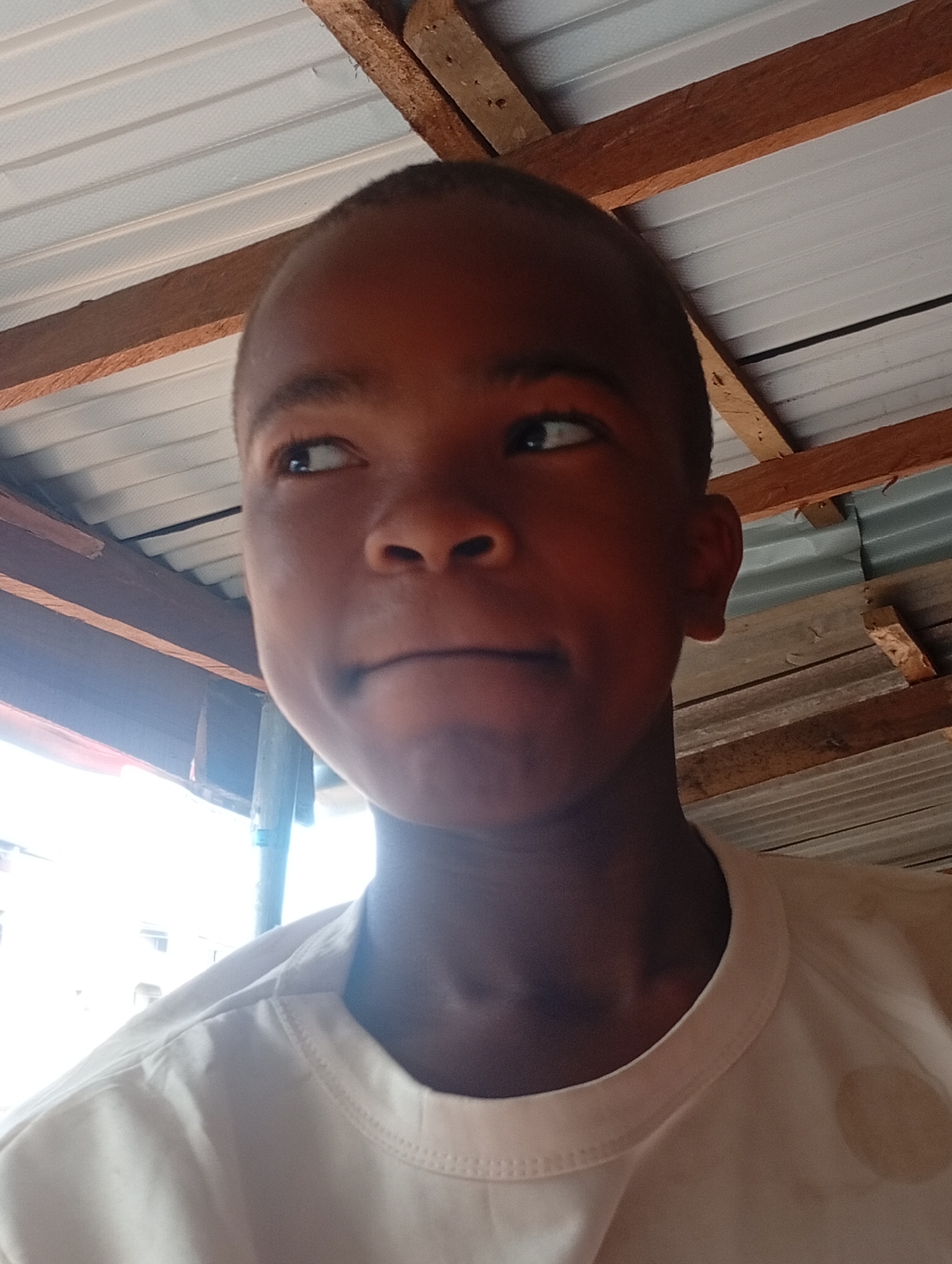 The image features a close-up portrait of a young person, likely a boy, with short, dark hair and dark skin. He is looking upwards and to his left (viewer's right) with a neutral expression, showing a slight hint of a smirk. His head is tilted slightly. He is wearing a light-colored, possibly white or off-white, crew-neck t-shirt.
The background primarily consists of an indoor ceiling structure. White or light grey corrugated metal sheets form the roof, supported by several visible wooden beams or planks that crisscross overhead. To the left side of the frame (viewer's right), a bright, overexposed area suggests an open window or doorway leading to the outside, indicating that it is daytime. Through this bright opening, faint outlines of distant structures or foliage can be discerned. The lighting is strong, particularly from the left.
No specific activity is taking place other than posing for the photograph. The setting appears to be a basic, perhaps utilitarian, indoor space. No visible text is present in the image. Given the context, this scene is located in Onitsha, Nigeria.