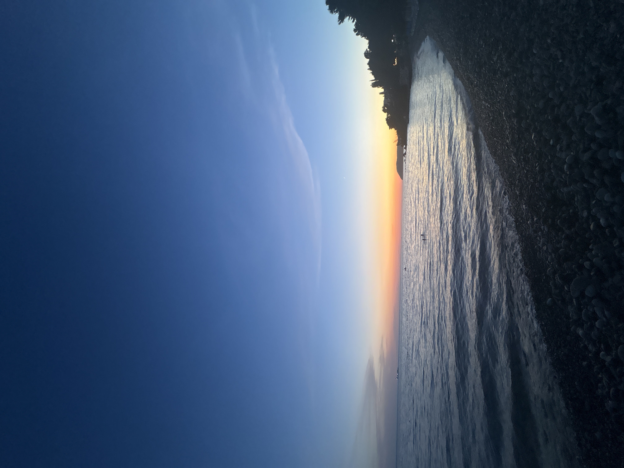 The image shows a pebble beach at sunset in Bar, Montenegro. The sky transitions from a deep blue at the top to a vibrant orange and yellow near the horizon. The sea is calm with gentle waves lapping the shore. In the distance, a ship can be seen on the water. A treeline with some structures is visible along the coast on the right side of the frame. There are a few people visible in the water. The foreground is dominated by dark-colored pebbles.