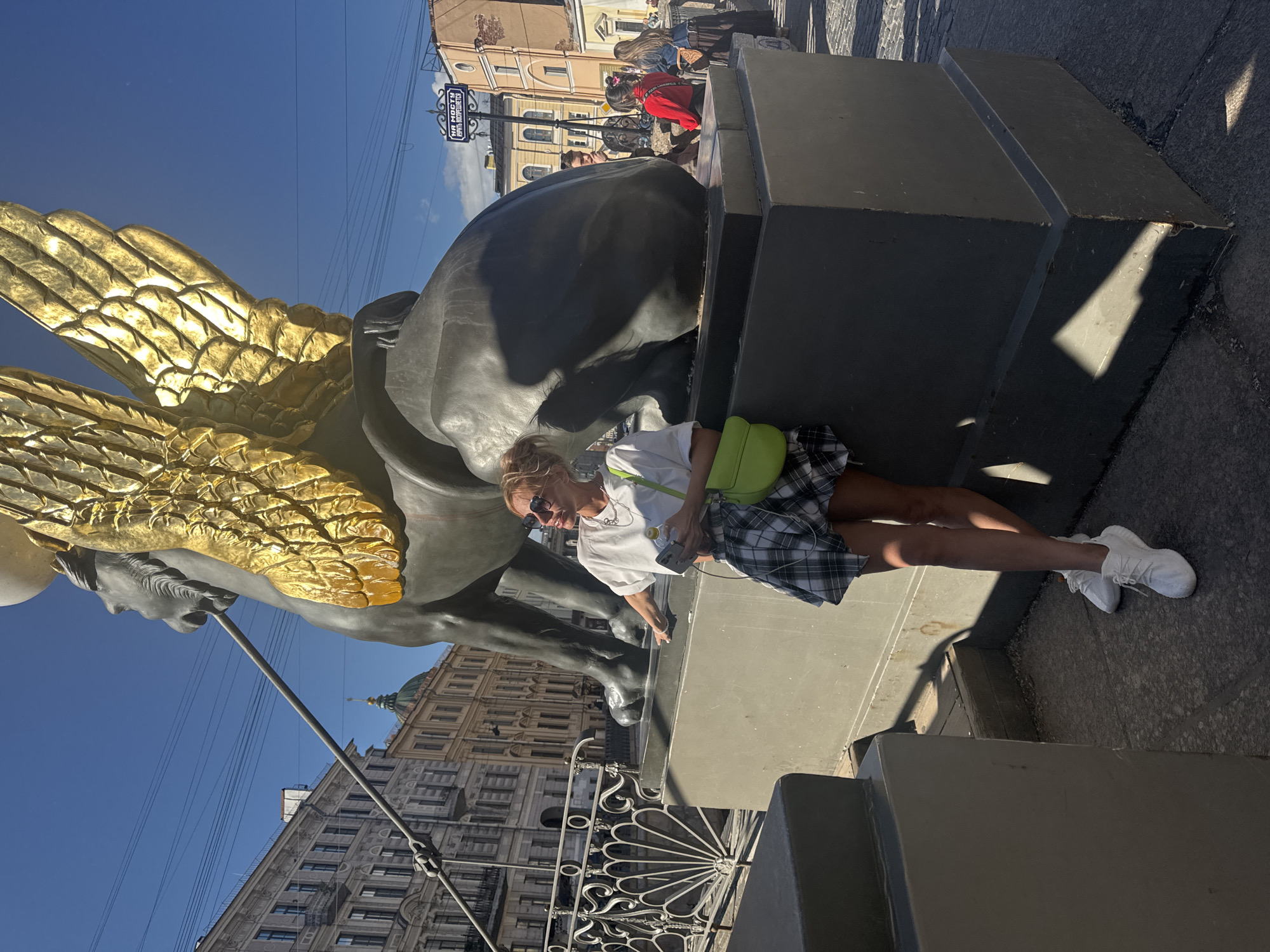 A young woman with blonde hair, wearing sunglasses, a white t-shirt, and a plaid skirt, is posing next to a large statue of a sphinx with golden wings. She is holding a bottle of water and a smartphone. The statue is dark gray and has an imposing presence. In the background, there are classic European-style buildings and a blue sky. The scene appears to be on a bridge, indicated by a metal railing in the foreground. A street sign in Russian is visible in the background. This is likely Saint Petersburg, Russia, given the architectural style and the presence of sphinx statues which are common decorative elements in the city, particularly on bridges over canals.