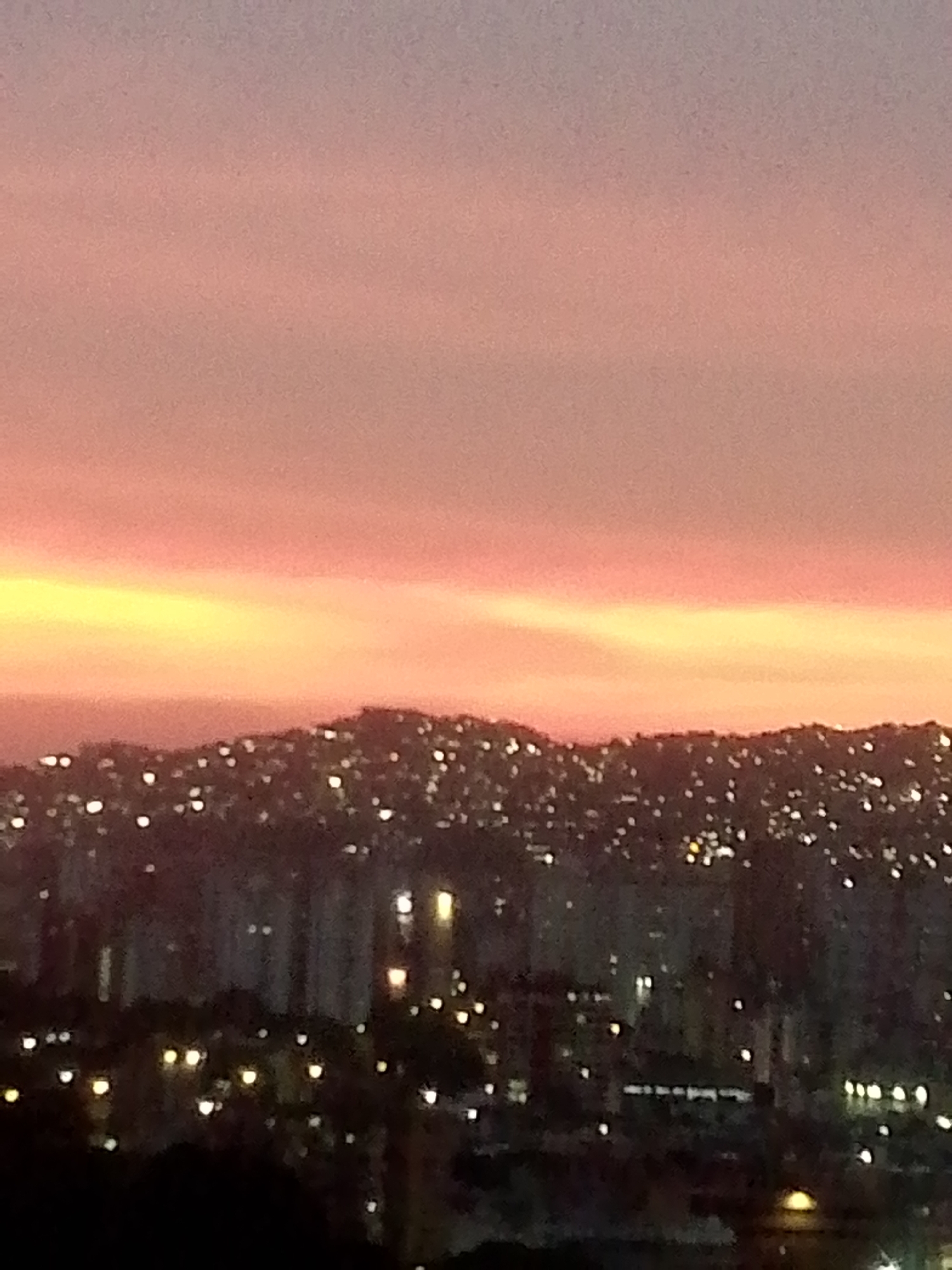 The image captures a cityscape under a sunset sky. The sky exhibits a gradient of colors, from pink at the top to orange near the horizon. Below the horizon is a dark landscape dotted with numerous small lights, indicative of a densely populated urban area. In the foreground, there are buildings and trees, also punctuated with lights.
Based on the urban landscape and distinctive hilly terrain visible in the distance, the scene is likely located in Caracas, Venezuela. The density of lights on the hillsides and in the valleys is characteristic of the city's urban sprawl.