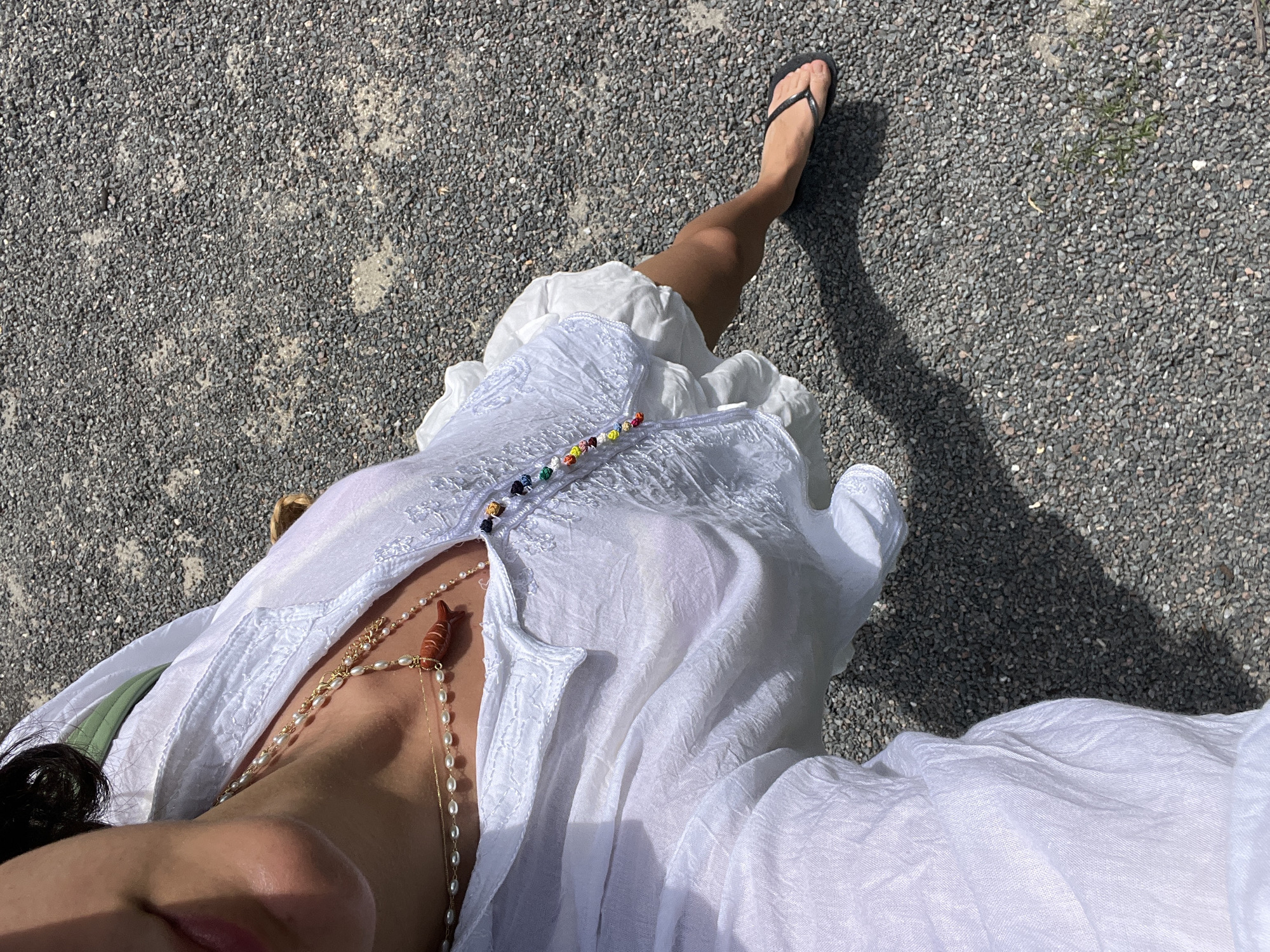 A high-angle shot depicts a person standing on a gravelly surface, likely a street or pathway. The individual is wearing a flowing white dress or garment with colorful buttons near the neckline. They also have several necklaces, including one with pearl-like beads and another with a lobster pendant. The person is wearing a black flip-flop on their right foot. The background consists of a gravelly surface, suggesting an outdoor setting. The lighting indicates a sunny day, and the shadow of the person's leg and foot is visible on the ground. The proximity of the shot makes it difficult to determine the specific location within Miami Beach, but the gravel surface suggests a park, beach access point, or residential area rather than the main tourist areas.