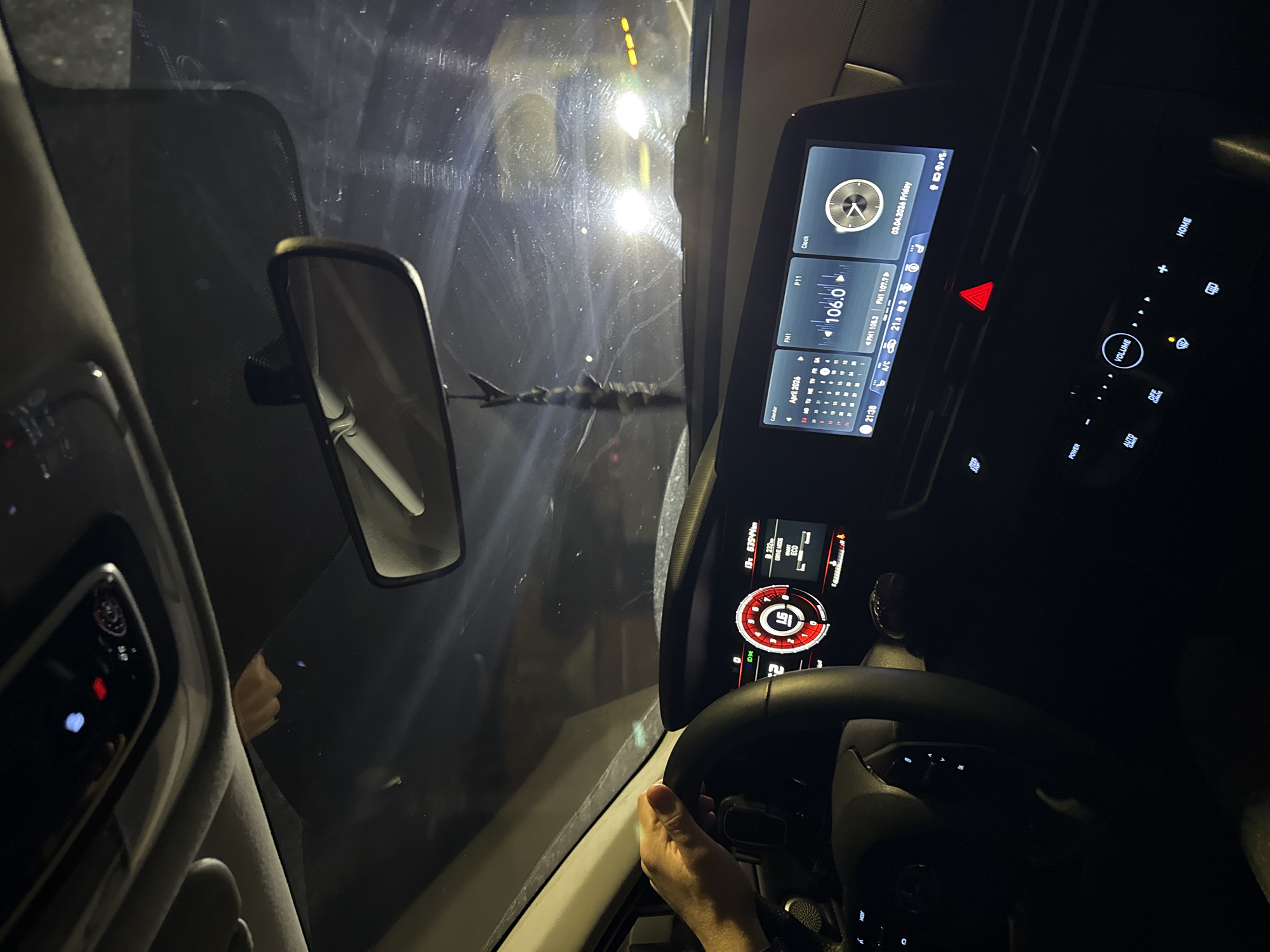 The image depicts the interior of a car at night. The windshield is visible with reflections of lights outside. Inside the car, the rear-view mirror, dashboard, and steering wheel are prominent. The dashboard displays various information including the time (21:38), date (April 3, 2026, Friday), radio station (FM 106.0), and a calendar. The speedometer indicates a speed of 16. The driver's hand is visible on the steering wheel. The driving mode is set to ECO. The vehicle has driven 63544 meters. The location and city are not identifiable.