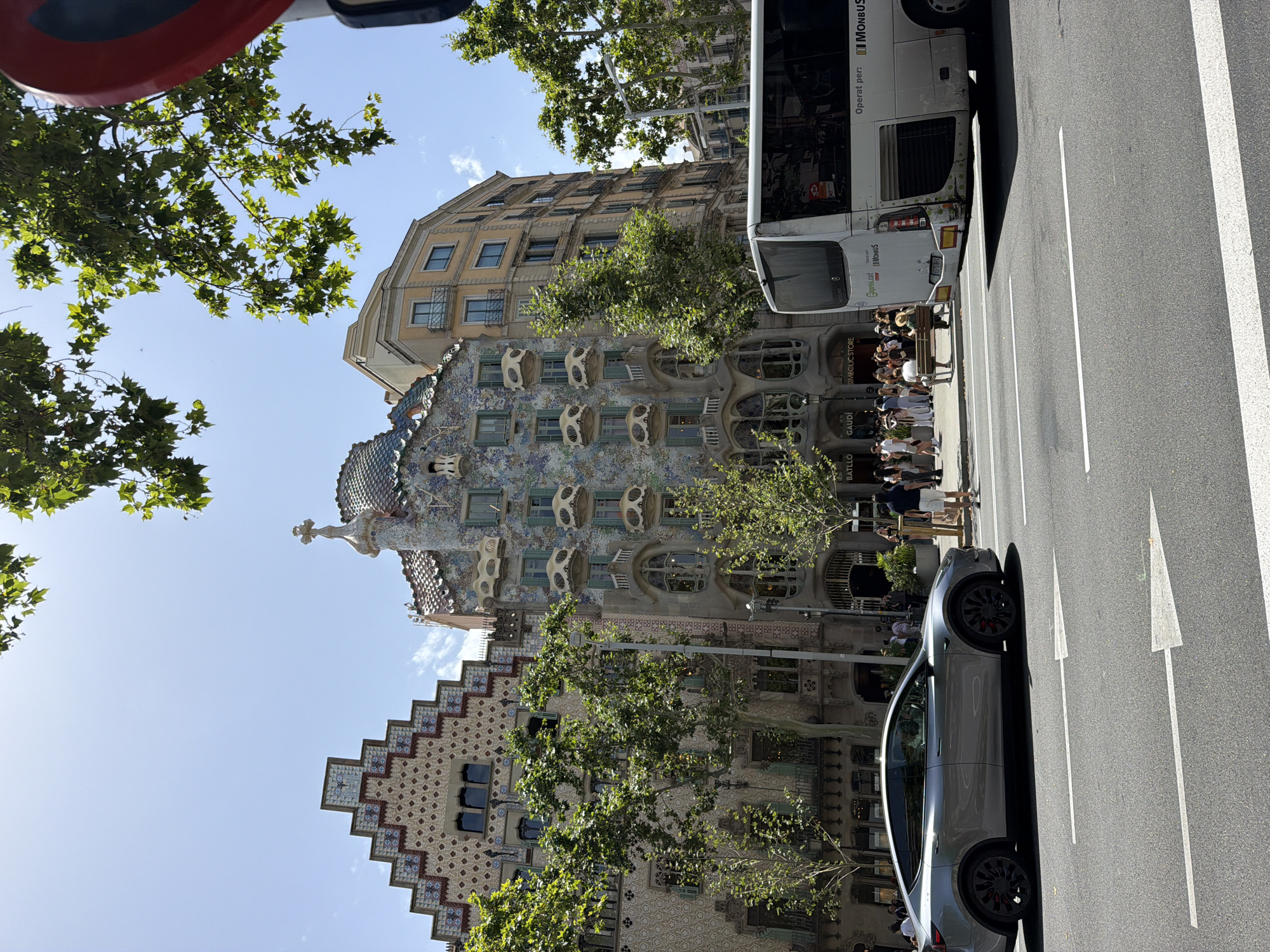 The image shows a street scene in Barcelona, Spain, featuring a distinctive building with a colorful, mosaic-tiled facade and organic, bone-like window frames. The building's architecture is ornate and appears to be a historical landmark. In front of the building, a group of people are gathered, possibly tourists, standing on a sidewalk. A modern grey electric car is parked on the street, aligned with white lane markings and an arrow indicating the direction of traffic. A white tour bus with text in Spanish and Catalan is also visible on the street, with "Operat per: MONBUS" and "Gaudí.cat" printed on its side. Trees with green foliage line the street, providing shade. The sky is blue with some scattered clouds, suggesting a clear, sunny day. The perspective of the photograph is from a low angle, looking upwards towards the buildings and sky. The scene captures a blend of historical architecture, modern transportation, and pedestrian activity in a lively urban environment.