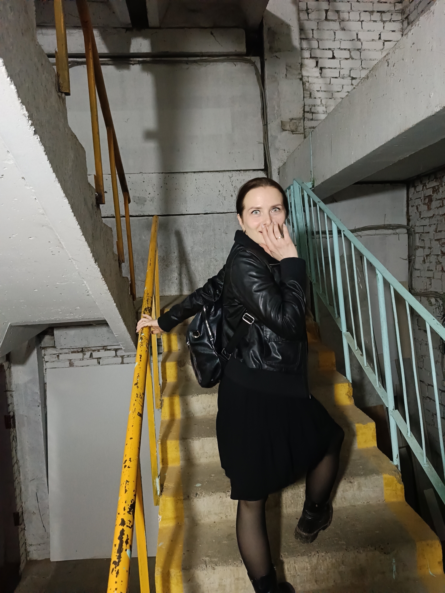 A woman is depicted ascending a concrete staircase in what appears to be an industrial or utilitarian building in Chaikovsky, Russia. She is caught in a moment of surprise or shock, with wide eyes and her left hand covering her mouth, as she looks directly towards the camera. Her right hand is resting on a yellow handrail as she steps up.
The woman is wearing a black leather jacket, a black skirt, black tights, and dark boots, with a black backpack slung over her shoulders. The staircase is made of concrete, with worn yellow safety stripes painted on the edges of the steps. Two distinct sets of handrails are visible: a prominent yellow one on the side the woman is touching, and a lighter blue-green handrail further up on the opposing side, indicating a switchback design.
The surrounding walls are a mix of raw concrete and white-painted brickwork, showing signs of age and wear. Exposed pipes and wiring run along the walls, contributing to the functional, unornamented aesthetic of the space. The lighting is artificial and appears somewhat dim, creating harsh shadows that emphasize the texture and structure of the environment. The overall atmosphere is stark and unadorned. No visible text is present in the image.