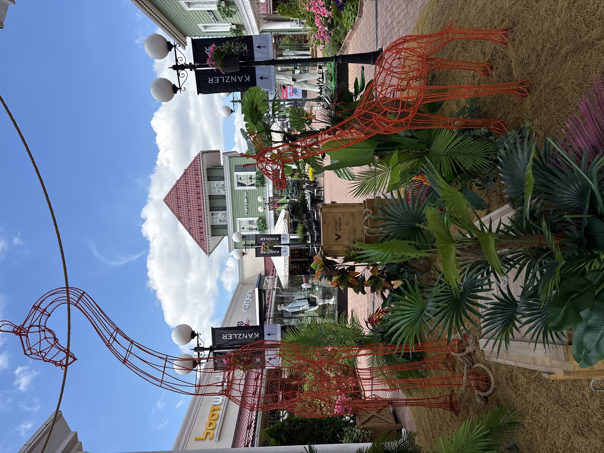 The image depicts an outdoor pedestrian shopping area. Two large, red wireframe giraffe sculptures are prominently displayed among lush green tropical plants and a sandy ground cover. In the background, a row of multi-story buildings with light green facades and red tiled roofs line the walkway. Signs for businesses such as "BOOTW", "Л'Этуаль", "KANZLER", and "COCCINELLE" are visible on the storefronts. Streetlights with white globe lamps are placed along the path, some adorned with black banners. A few tables and chairs are set up outside a restaurant. The sky is blue with scattered white clouds.