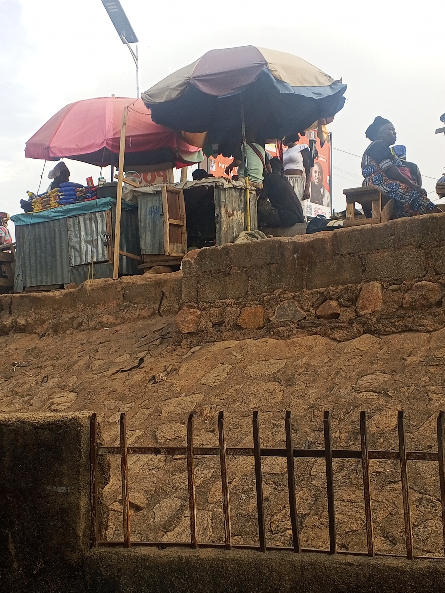 The image depicts a street scene in Gwong, Nigeria. A rusty metal railing stands at the bottom of the frame, partially obscuring a textured, rocky ground surface. Above this, a retaining wall built from concrete blocks and stones supports a higher level where commercial activity is taking place. Two makeshift stalls, constructed from corrugated metal and wood, are visible. These stalls are shaded by large umbrellas, one pink and the other blue and beige. People are clustered around the stalls, some standing and others sitting. One woman sits on a wooden stool, wearing a patterned dress. A political poster is partially visible in the background. A solar-powered street light rises above the scene.