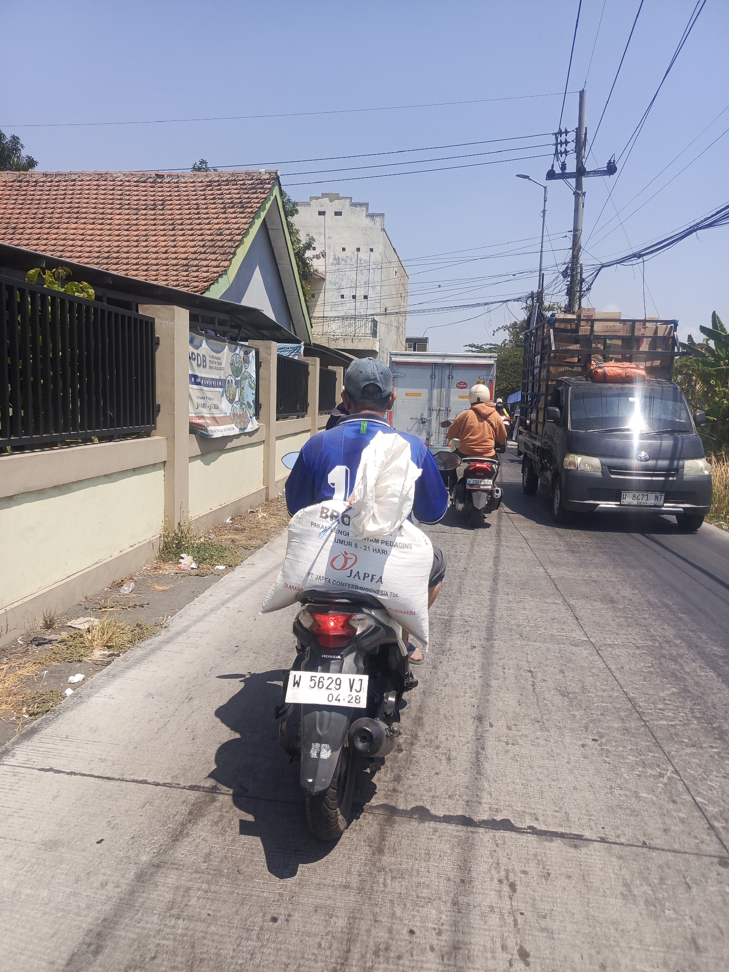 In Banjarsari, Indonesia, on a clear, sunny day, a person rides a motorcycle on a paved road. The motorcycle is carrying a large, white bag tied to the back, with text visible on it. The rider is wearing a blue shirt with a white number "1" on the back and a gray cap. In front of them, another motorcycle is visible with a rider wearing an orange jacket and a white helmet. Further ahead, a black truck with wooden racks filled with boxes is driving on the same road. The environment includes buildings with terracotta roofs and white walls, a black fence, utility poles with numerous wires, and some sparse vegetation along the roadside. The road surface shows signs of wear and some oil stains. The overall scene depicts a typical day of transportation and commerce in the area.