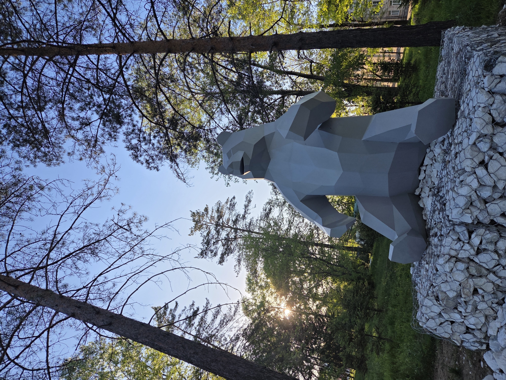 The image captures a low-angle perspective of a polygonal, gray bear sculpture set against a backdrop of trees and a clear sky. The bear is positioned atop a gabion filled with white stones. The sunlight filters through the foliage, creating a flare effect. The sculpture is located outdoors, in what appears to be a park or garden. Based on the provided information, the location is Birobidzhan, Russia.