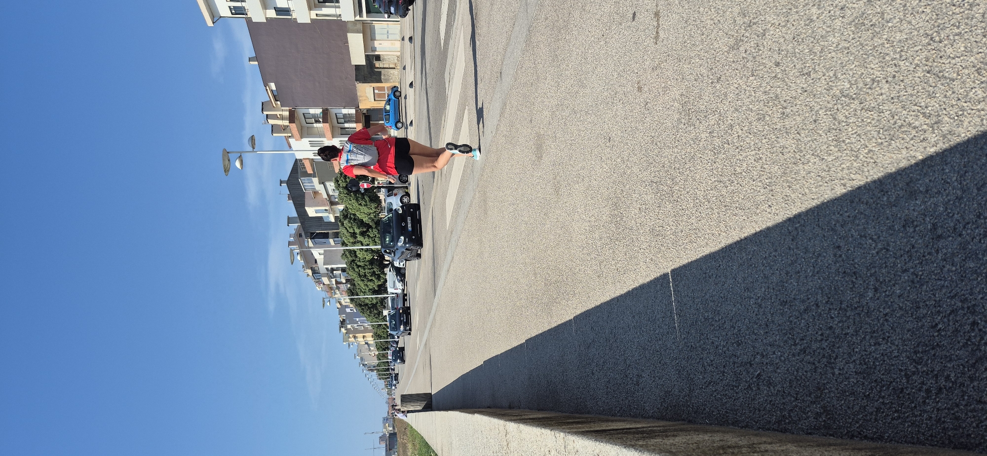 The image shows a person running on a sidewalk next to a street with traffic. The scene is located in Vila do Conde, Portugal.
**Detailed Description:**
**Location:** The image depicts a street scene in Vila do Conde, Portugal, likely along the coastline given the presence of a seawall and the distant open sky. The architecture of the buildings suggests a European coastal town.
**People:**
* A person, viewed from behind, is running on a paved pedestrian walkway that runs parallel to the street. This individual is wearing a red t-shirt, black shorts, and a light-colored backpack. They have athletic footwear on.
**Objects and Environment:**
* **Road and Traffic:** A paved road with multiple lanes is visible, with several cars in motion. One black smart car is prominently featured in the mid-ground. A blue car is parked further down the street. A pedestrian crossing with white stripes is in the foreground.
* **Buildings:** A row of multi-story buildings with various architectural styles lines the street on the right side of the image. They appear to be residential or commercial properties.
* **Street Furniture:** Streetlights with a distinctive curved design are spaced along the side of the road, indicating it's a public urban area.
* **Seawall:** A low concrete seawall is present on the left side of the image, separating the pedestrian walkway from what is likely a beach or the sea.
* **Vegetation:** Trees with lush green foliage are visible between the road and the buildings, adding a touch of nature to the urban landscape.
* **Sky:** The sky is a clear, bright blue, suggesting a sunny day with good weather.
* **Shadows:** A distinct shadow is cast across the paved walkway from the seawall, indicating the position of the sun.
**Action:** The primary action depicted is the person running on the walkway. The cars on the road are in motion, indicating typical street activity.
**Interactions:** There are no direct interactions between the people in the image. The runner is engaged in their activity, and the vehicles are moving independently.
**Overall Impression:** The image captures a moment of daily life in a Portuguese coastal city, with a focus on an individual exercising in an urban environment. The clear weather and open sky contribute to a sense of a pleasant day.