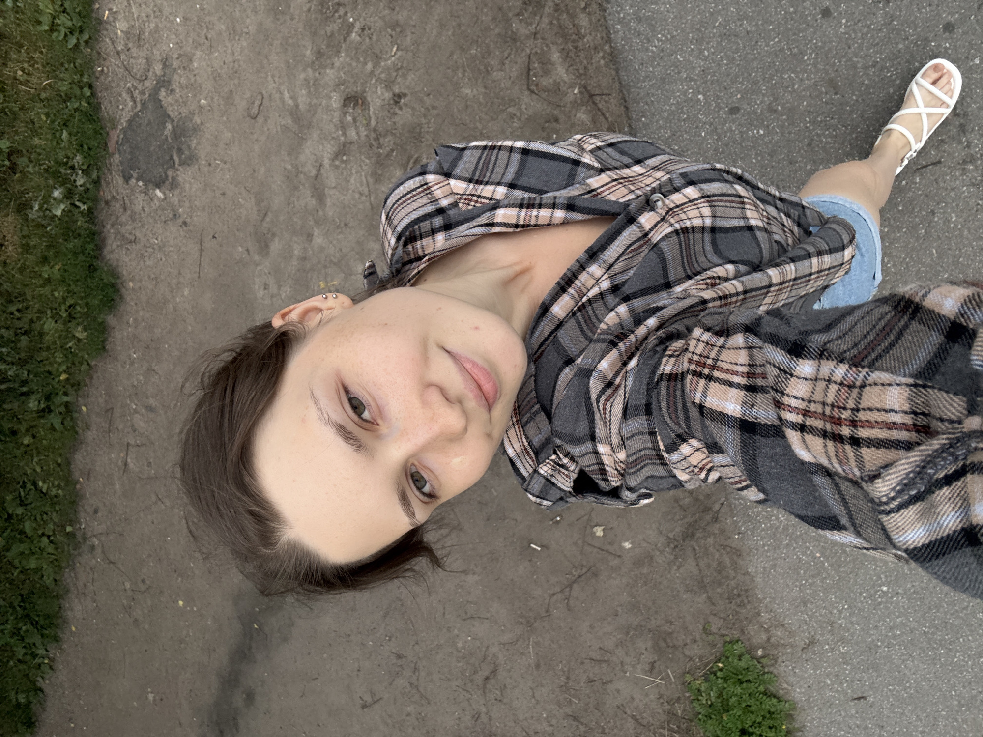 The media file is a selfie taken from a high angle, showing a young woman looking up at the camera. She is wearing a plaid shirt and denim shorts, with white sandals on her feet. The woman has light brown hair and a fair complexion with freckles. She is smiling softly, and her expression conveys a sense of ease and happiness.
The setting appears to be an outdoor environment, possibly a street or a park, with concrete ground and some patches of grass. There are some fallen leaves and small debris scattered on the ground, suggesting it might be autumn. The lighting is natural, indicating daytime, but the overall tone of the image is somewhat subdued, possibly due to overcast weather.
The background shows a typical urban or suburban street scene, with a slightly worn concrete surface. The presence of cigarette butts on the ground suggests a public area. The woman's attire and the casual setting suggest an everyday moment, perhaps a stroll or a break outdoors. The selfie format and her direct gaze create an intimate connection with the viewer. There is no visible text in the image. The context provided mentions Saint Petersburg, Russia, which may inform the visual cues, such as the type of pavement or vegetation.