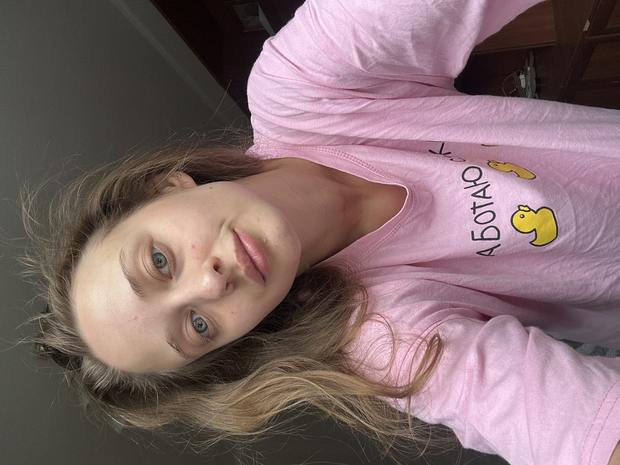 A young woman with light brown hair and blue eyes is shown in a selfie. She is wearing a pink long-sleeved t-shirt with yellow duck illustrations and some Cyrillic text. The text visible on the shirt appears to be "АБОТАЮ", which translates to "working" in Russian. The woman has a neutral expression on her face. The background is dark and appears to be indoors, with a hint of wooden furniture and a window with natural light coming through. The overall impression is that of a casual, candid moment captured at home.