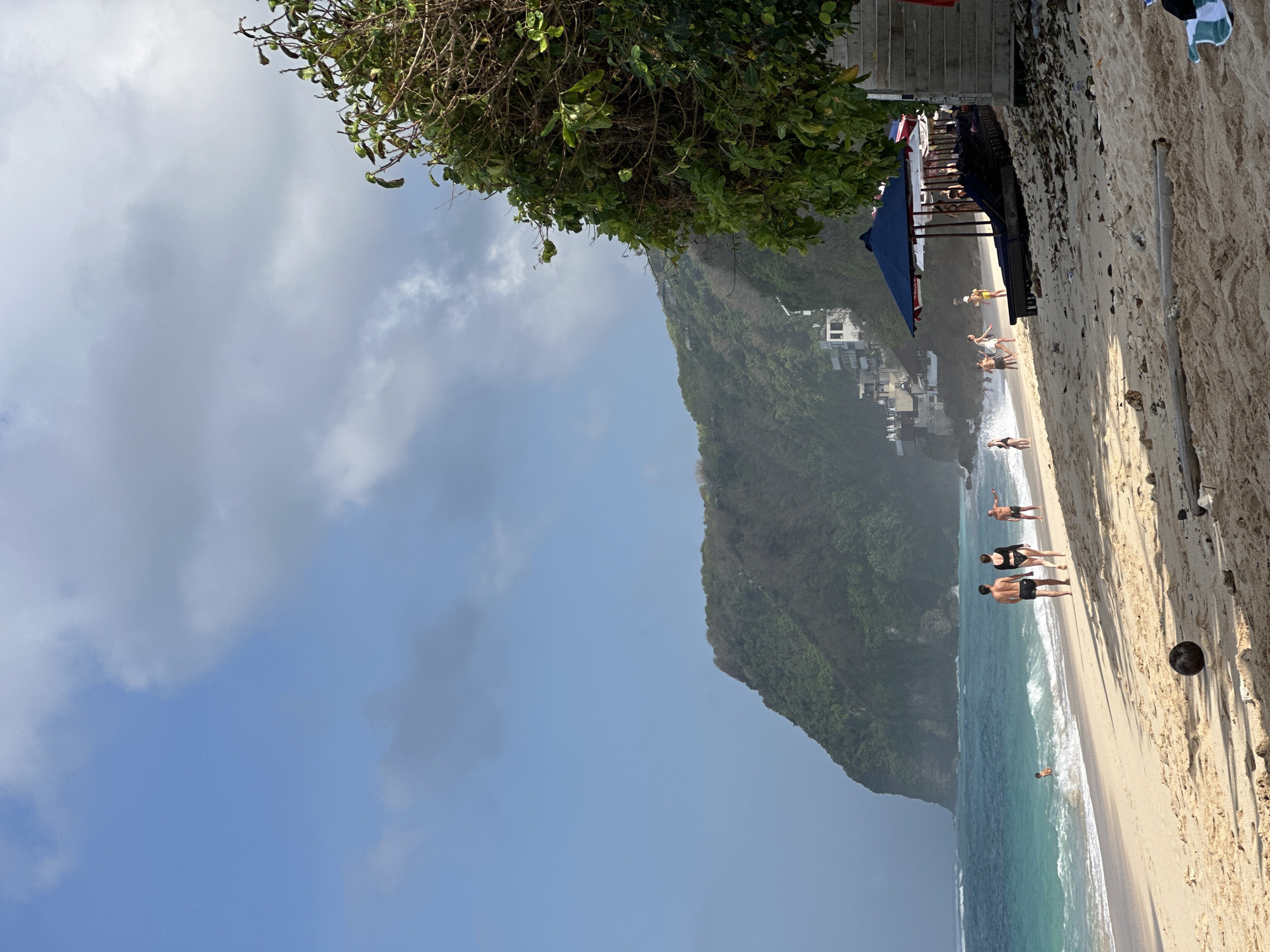 This image depicts a tropical beach scene in Kuta Selatan, Indonesia. In the foreground, a sandy beach is littered with debris, including driftwood and a coconut. In the middle ground, several people are walking along the shoreline and swimming in the turquoise ocean. Some are dressed in swimwear, while others are in casual beach attire. A cluster of lounge chairs and umbrellas are set up on the sand, suggesting a resort or beach club area. In the background, a lush green cliff face rises dramatically from the sea, dotted with buildings. The sky is a mix of blue sky and scattered clouds, and the lighting suggests it is daytime, possibly late afternoon given the soft sunlight and shadows. The overall atmosphere is one of a serene and active beach day.