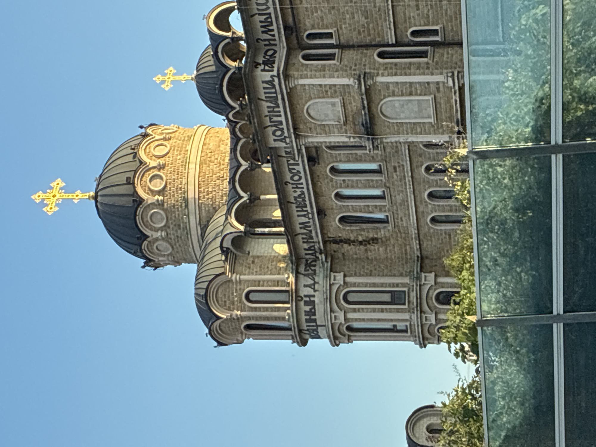 The image shows the exterior of the Church of the Resurrection of Christ in Sirius, Russia. The church is constructed of light-colored stone and features multiple domes, the largest of which is topped with a golden cross. The architecture is ornate, with arched windows and decorative stonework. Cyrillic inscriptions are visible on the facade of the building. Green trees are seen in the foreground, partially obscuring a glass barrier. The sky is clear and blue.