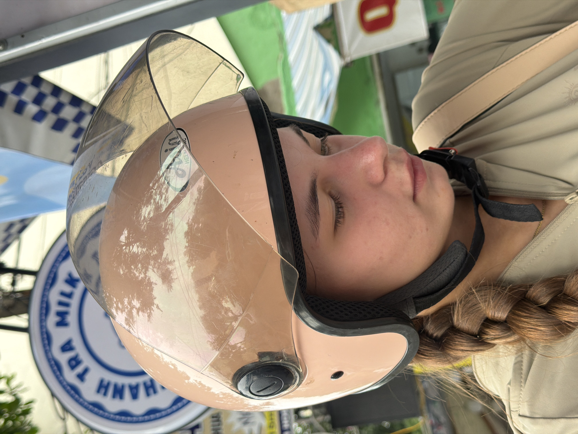 The image shows a close-up of a young woman wearing a light pink helmet with a clear visor. She has a braid of brown hair falling over her shoulder. She is wearing a light-colored jacket with a strap from a bag visible on her shoulder. The background suggests an outdoor setting, possibly a street scene in Nha Trang, Vietnam, with a blurred sign that reads "HANH TRA MILK" in blue lettering on a white circular background. Other blurred signs and awnings are also visible, indicating a commercial or street-side location. The overall impression is of a casual, everyday moment captured outdoors.