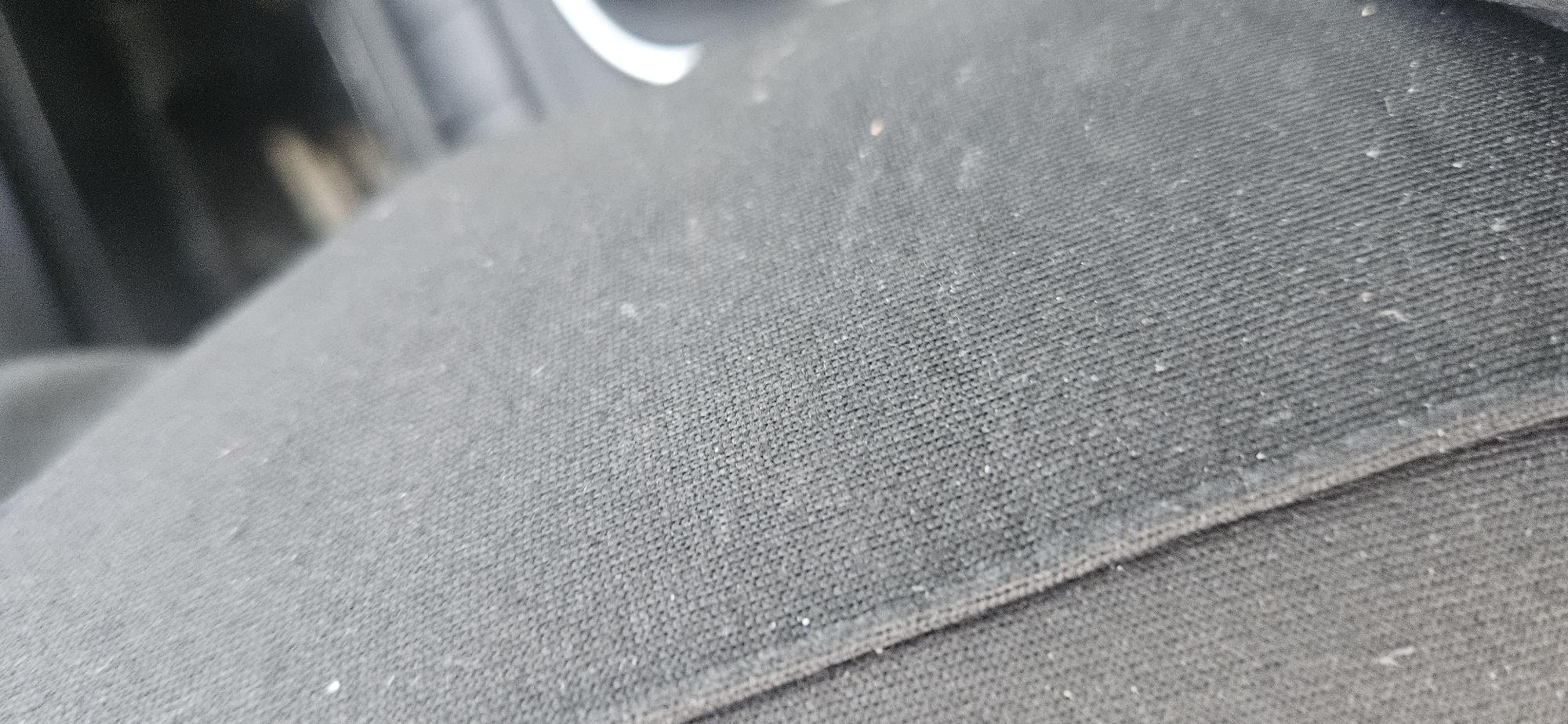 The image presents a close-up, angled view of a black, textured fabric surface, most likely a car seat or a similar upholstered interior component. The fabric exhibits a fine, woven pattern, and its surface is visibly covered with an accumulation of dust, lint, and small, light-colored particles, suggesting a lack of recent cleaning. A seam runs diagonally through the lower right section of the frame, delineating two distinct areas of the fabric.
The background is heavily blurred and out of focus, showing indistinct dark shapes and a small, prominent white circular object in the upper right. This white object, along with the overall context, further suggests an interior setting, likely within a vehicle. No people are present, and no specific activity is taking place. The lighting appears even and neutral, consistent with an interior environment. There is no visible text in the image. While the location context is given as Plovdiv, Bulgaria, this information is not visually discernible from the content of the image.