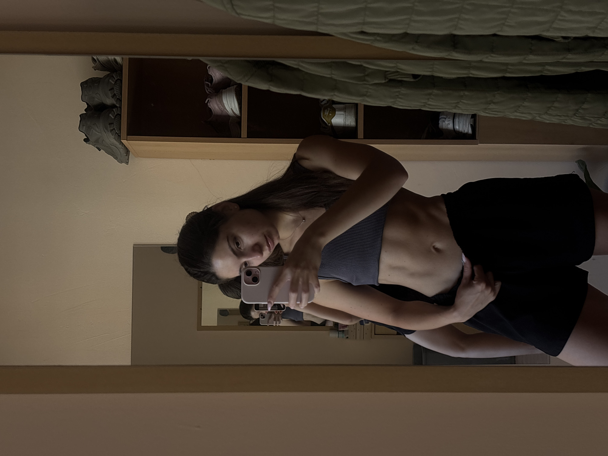 A young woman with dark hair tied back is taking a selfie in a mirror. She is wearing a dark grey ribbed tank top and black shorts. Her midriff is visible, showing well-defined abdominal muscles. She is holding a pink iPhone in her right hand and has her left hand on her shorts. In the background, reflected in the mirror, is another mirror and a shelf unit filled with shoes and other items. A light green quilted jacket is hanging to the right of the mirror. The overall setting appears to be a domestic interior, possibly a bedroom or dressing area. There is no indication of the city of Varna, Bulgaria.