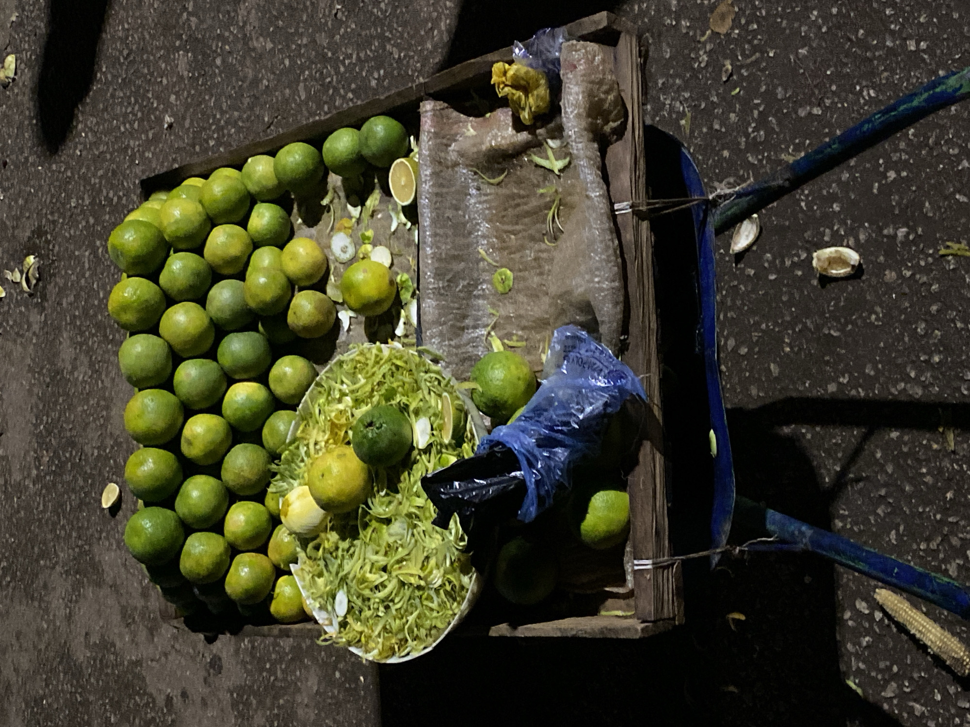 An overhead shot depicts a vendor's wooden pushcart or stall, likely situated on a street or market in Bukuru, Nigeria, during the night or in very low light conditions. The cart rests on a dark, granular paved surface, possibly asphalt.
The main subjects are numerous round, green and yellowish-green citrus fruits, resembling unripe oranges or limes. A large quantity of these fruits is piled on the left side of the cart. In the center, a white, shallow bowl or plate is overflowing with a substantial mound of finely shredded green peelings, indicating active fruit preparation, possibly for juice or a snack. Several whole fruits and discarded peels are scattered around this central pile.
On the right side of the cart, more fruits are visible. Some are contained within a translucent blue plastic bag, where the partial text "POLY" can be faintly seen in white. A crumpled yellow plastic bag is also present. The cart itself has a blue metal handle extending towards the right. Various fruit peels, scraps, and a small corn cob or husk are scattered on the ground around the cart, reinforcing the idea of an active vendor operation. The deep shadows and dark background strongly suggest the image was taken at night under artificial illumination. No individuals are visible in the frame.