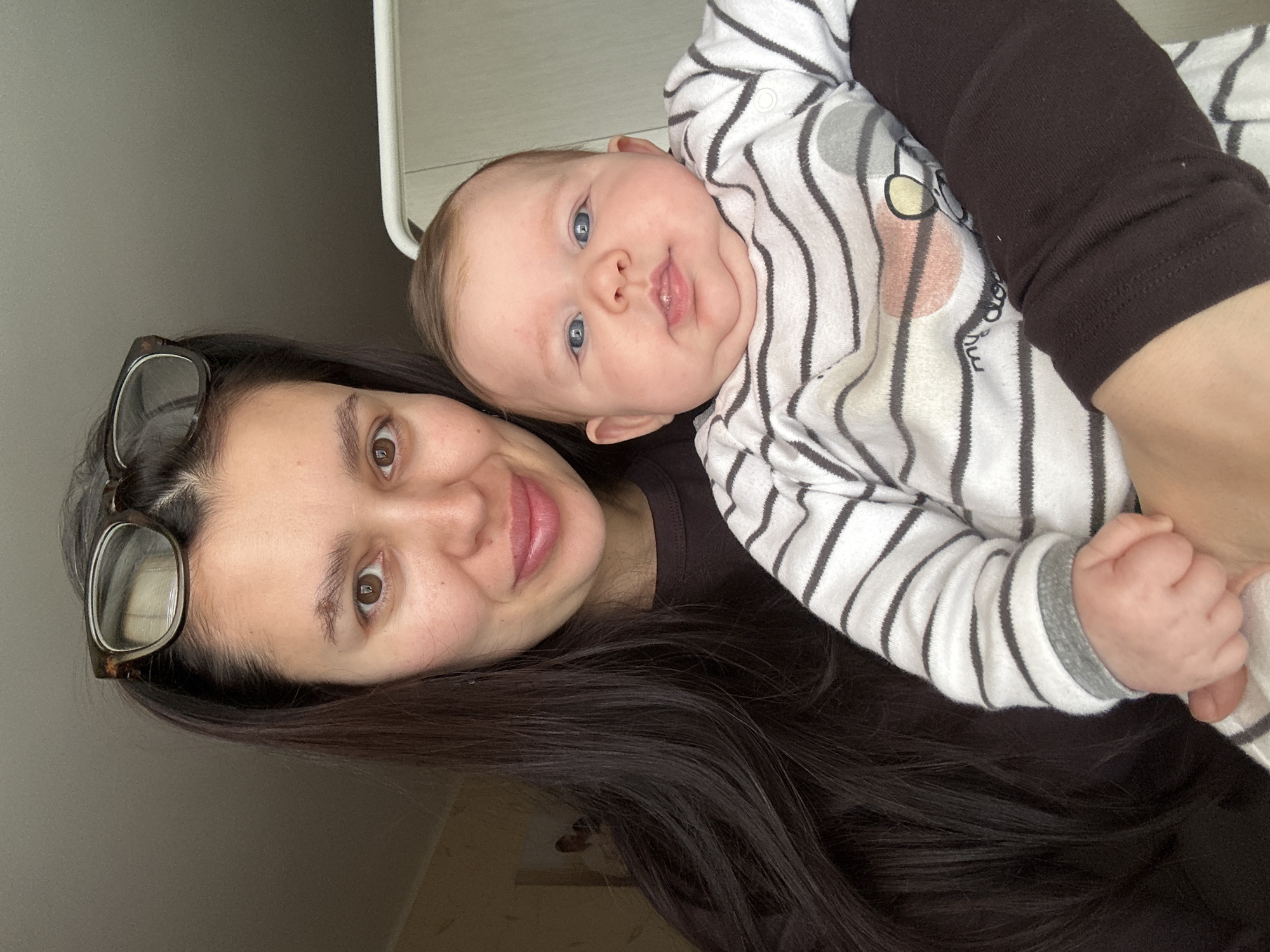 This intimate portrait captures a mother and her baby indoors, likely in Chaikovsky, Russia. The image shows the two subjects looking directly at the camera with gentle expressions.
On the left, the woman, who appears to be the mother, has long, dark brown hair framing her face. She has warm brown eyes and a soft smile. A pair of tortoiseshell-framed glasses are pushed up onto her head, resting on her hair. She is wearing a dark, possibly brown, long-sleeved top. Her skin is fair with visible freckles.
Cradled in her arms, and positioned towards the right side of the frame, is an infant. The baby has captivating blue eyes and light, fine hair, with a healthy, rounded face. The baby is dressed in a white and grey striped long-sleeved top or onesie, which also features a subtle, light-colored design on the chest area. The baby has a slight, open-mouthed smile, conveying a sense of contentment.
The background is a simple, plain light grey surface, suggesting an uncluttered indoor setting, possibly a home. The lighting is soft and even, typical of diffused natural light indoors during the daytime. Both mother and child exhibit a calm and happy demeanor, capturing a tender moment of connection. There is no visible text within the image.