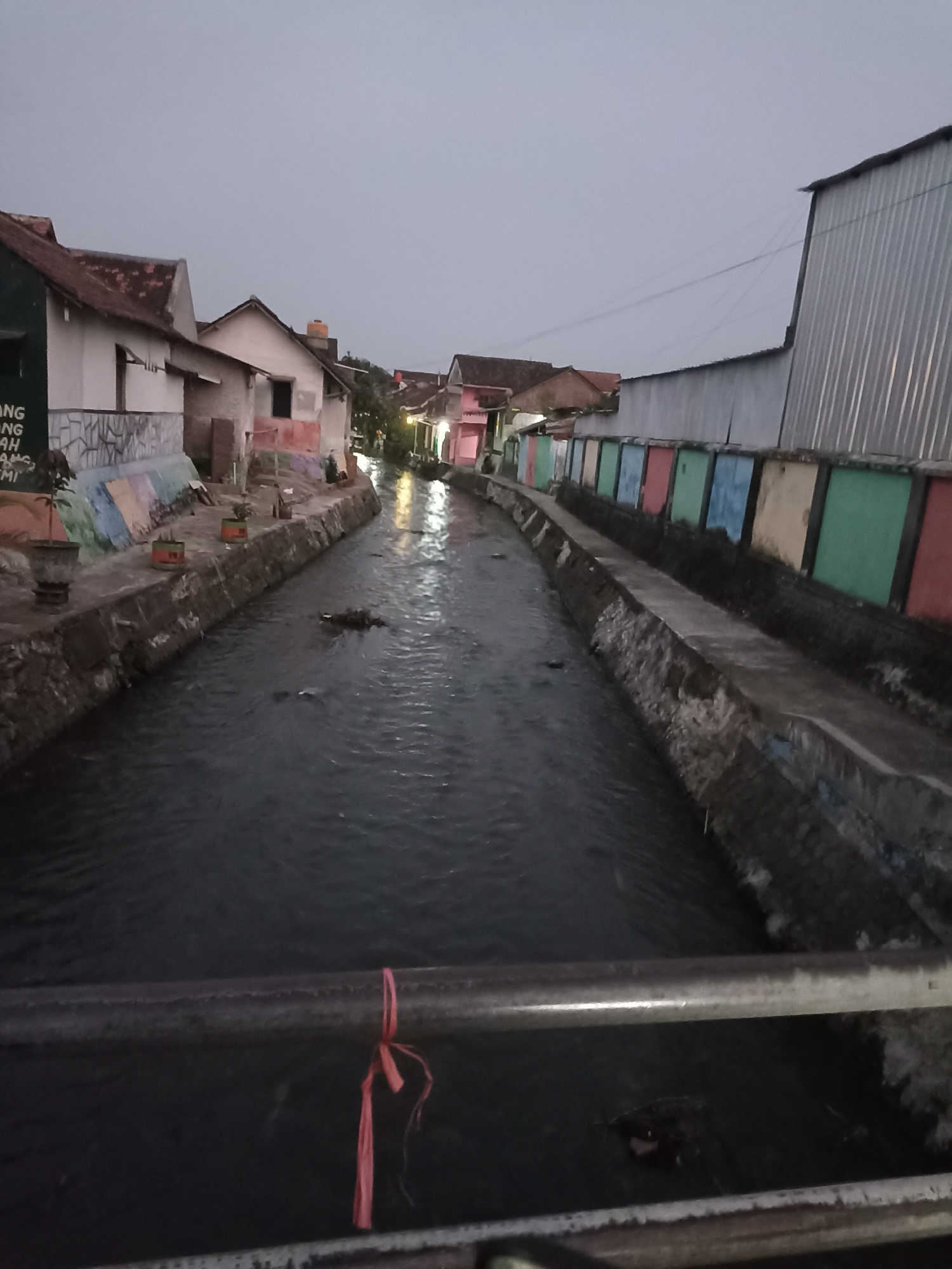 In Ditotrunan, Indonesia, a dimly lit scene depicts a narrow canal flowing between two rows of buildings. The water in the canal is dark and appears to be somewhat polluted. On the left bank, a wall is adorned with colorful murals and planters with small trees. Several houses are visible behind this wall, featuring red-tiled roofs and light-colored facades. Some of these houses have visible windows and doorways. On the right bank, a long wall is painted with brightly colored rectangular panels in shades of pink, green, yellow, and blue. Beyond this wall, a large building with a corrugated metal roof is prominent. A metal railing with a red tie is in the foreground, partially obscuring the view of the canal. The sky above is overcast and gray, suggesting it is either early morning or late evening. The overall impression is of a residential area with a prominent water channel.