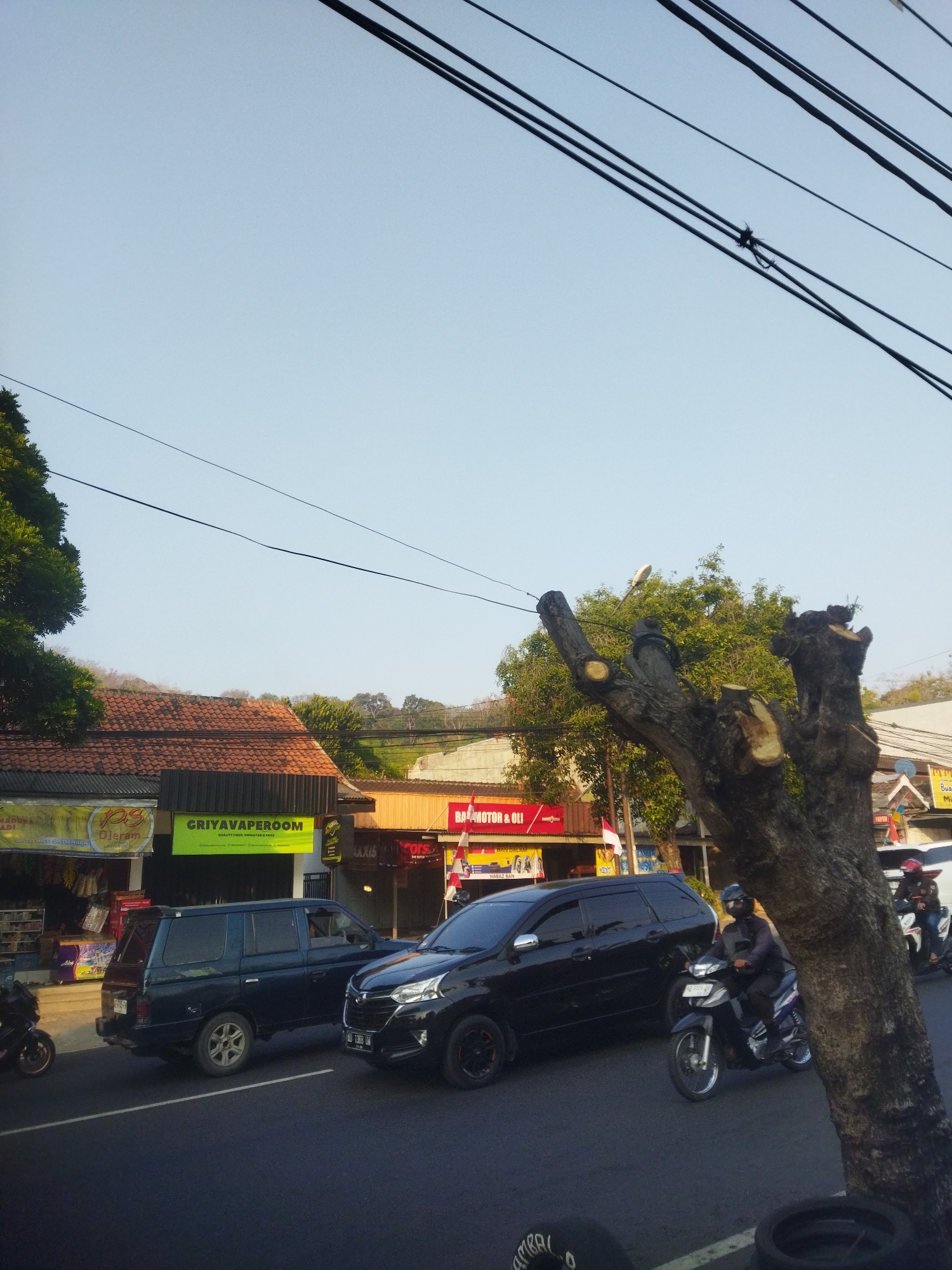 The image depicts a street scene in Wonogiri, Indonesia. The sky is clear and blue, with several overhead power lines crossing the frame. On the left side of the street, a row of buildings lines the road. One building has a sign that reads "GRIYAVAPEROOM" in large green letters. In front of this building, a dark blue SUV is parked. A black car is driving on the road in front of the SUV. A motorcyclist wearing a dark jacket and helmet is riding a motorcycle in the foreground, moving from left to right. To the right of the road, a large tree trunk with several cut branches is prominently featured. In the background, more buildings and trees are visible, with a hillside in the far distance. The overall scene suggests a typical day in a populated area, with traffic and commercial establishments.