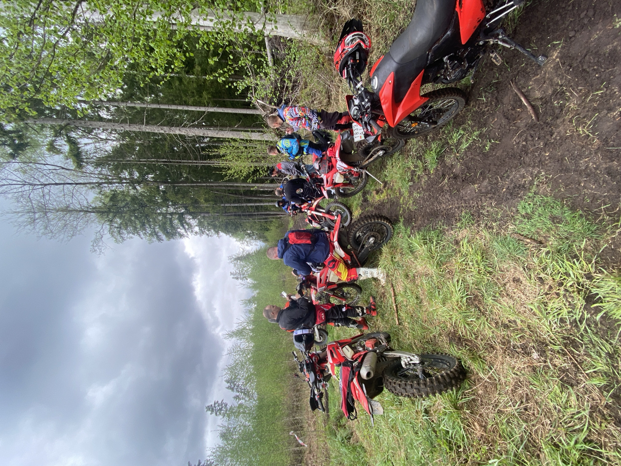 A group of people on dirt bikes are stopped on a grassy area alongside a forest edge in Гожскі сельскі Савет, Belarus. The bikes are primarily red and black, and the riders are wearing various riding gear. Some of the riders are standing near their bikes, while others are seated. The sky is overcast, and the ground appears damp, suggesting recent rain.