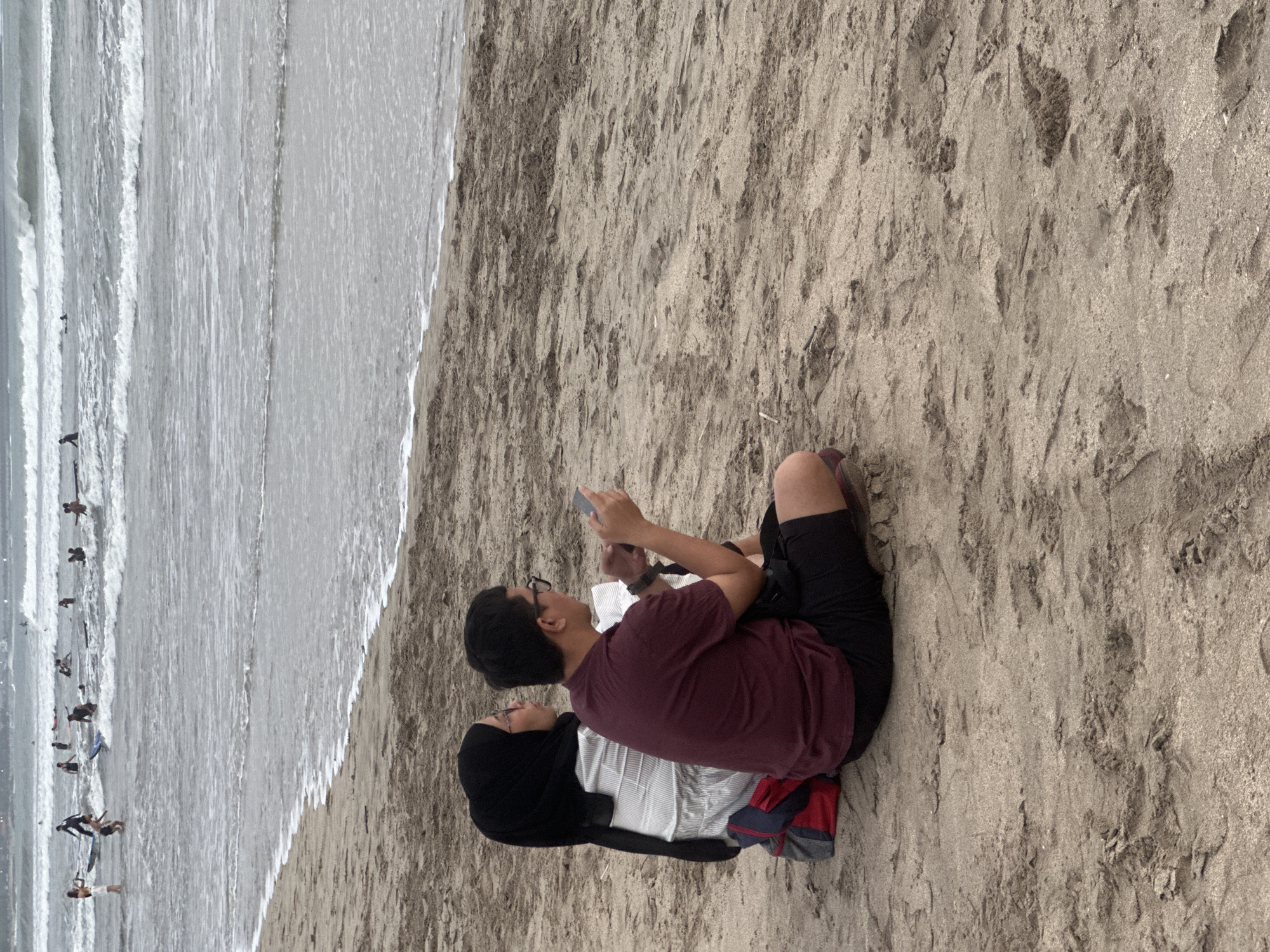 A medium shot shows two individuals, a woman and a man, sitting on a sandy beach facing away from the camera, towards the ocean. The woman is on the left, wearing a black hijab and a white top. The man is on the right, wearing a maroon t-shirt and black shorts. He is holding a mobile phone in his hands, and both individuals appear engaged with their devices or observing something in the distance. A backpack with red and blue accents is placed behind the couple. The ocean stretches out behind them with waves and several people can be seen in the water, some with surfboards. The sand in the foreground is textured with footprints and debris.