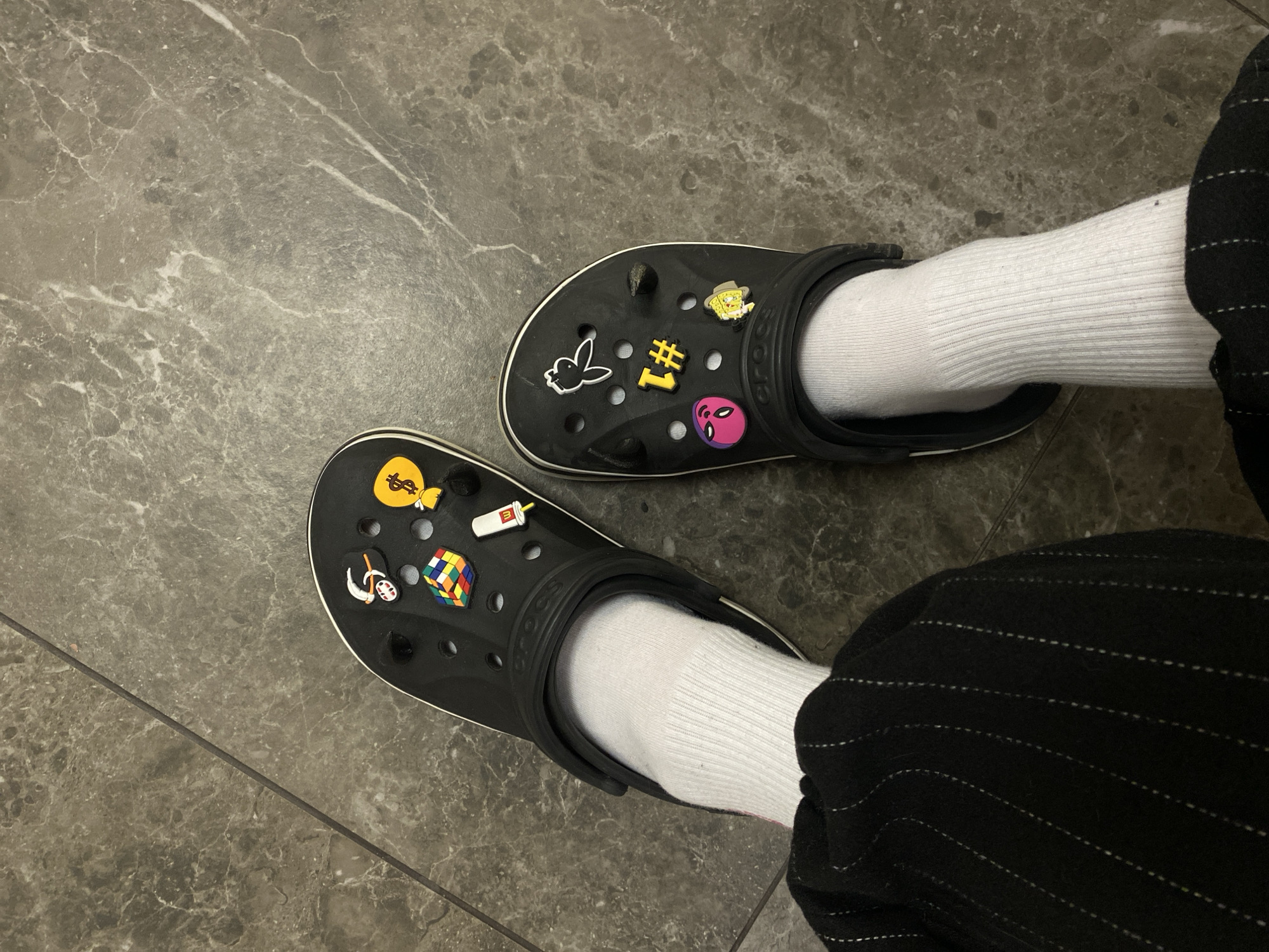 The image shows two feet wearing black Crocs and white socks, standing on a dark gray tiled floor. The right foot is partially obscured by black pinstriped trousers. The Crocs are adorned with various Jibbitz charms, including a dollar sign bag, a Rubik's cube, a soda cup, a ghost figure, a Playboy bunny, a "Number 1" symbol, an alien head, and Spongebob Squarepants. The tiles appear to be arranged in a linear pattern. The location is not discernible from the provided image and cannot be identified as Votkinsk, Russia.