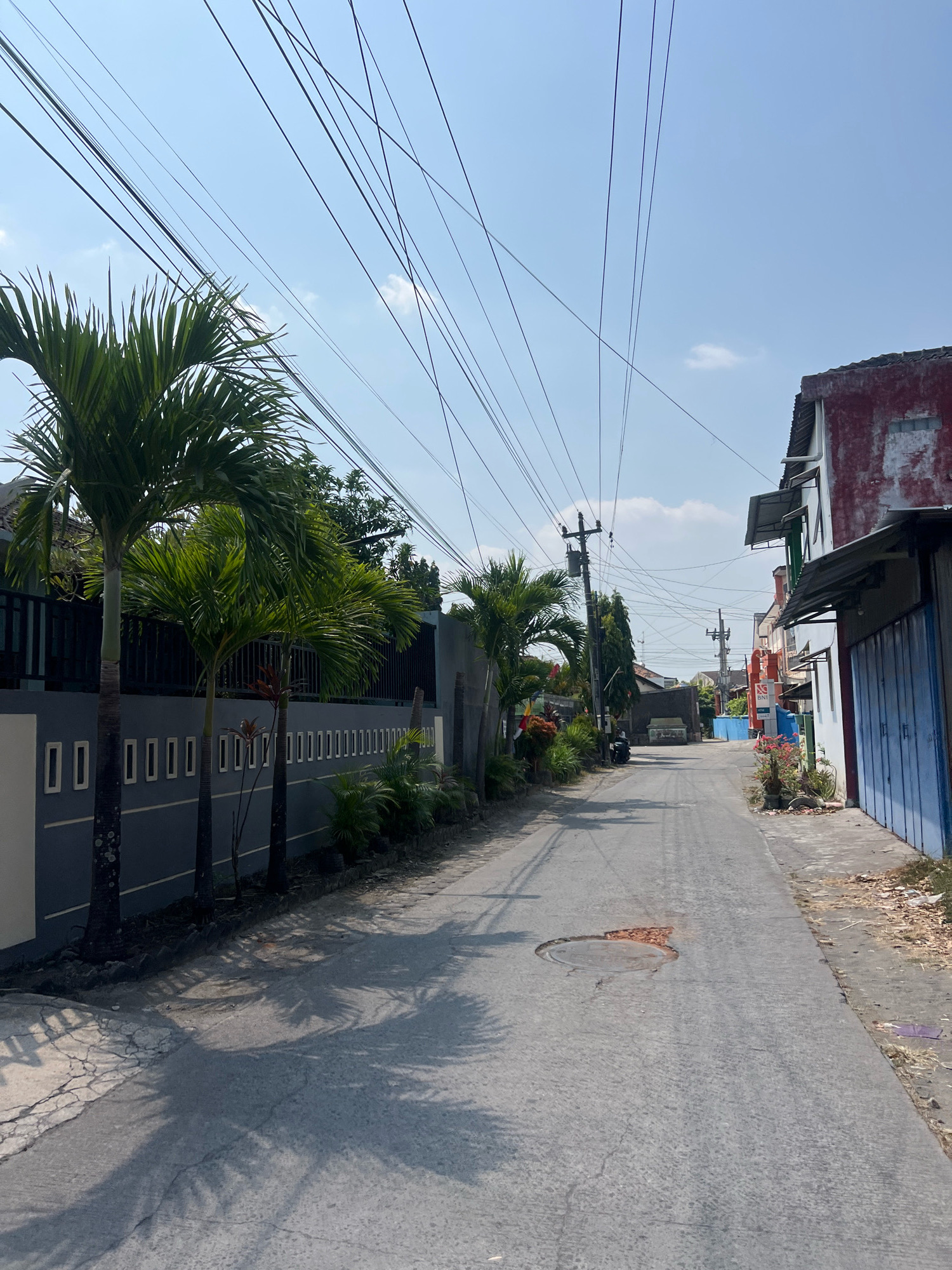 The image depicts a narrow, paved street in Sewon, Indonesia, under a clear blue sky with scattered clouds. Power lines crisscross the sky, supported by utility poles. On the left side of the street, a low, gray wall with rectangular indentations runs alongside several palm trees. Further down the street, buildings with red and white facades are visible, some with corrugated metal awnings and blue metal doors. A motorcycle is parked on the left side of the road. The pavement shows signs of wear, with a circular manhole cover in the center of the road that appears to be damaged, exposing reddish-brown material. Shadows of palm fronds are cast across the road and the wall. The overall impression is a residential street in a tropical climate.
