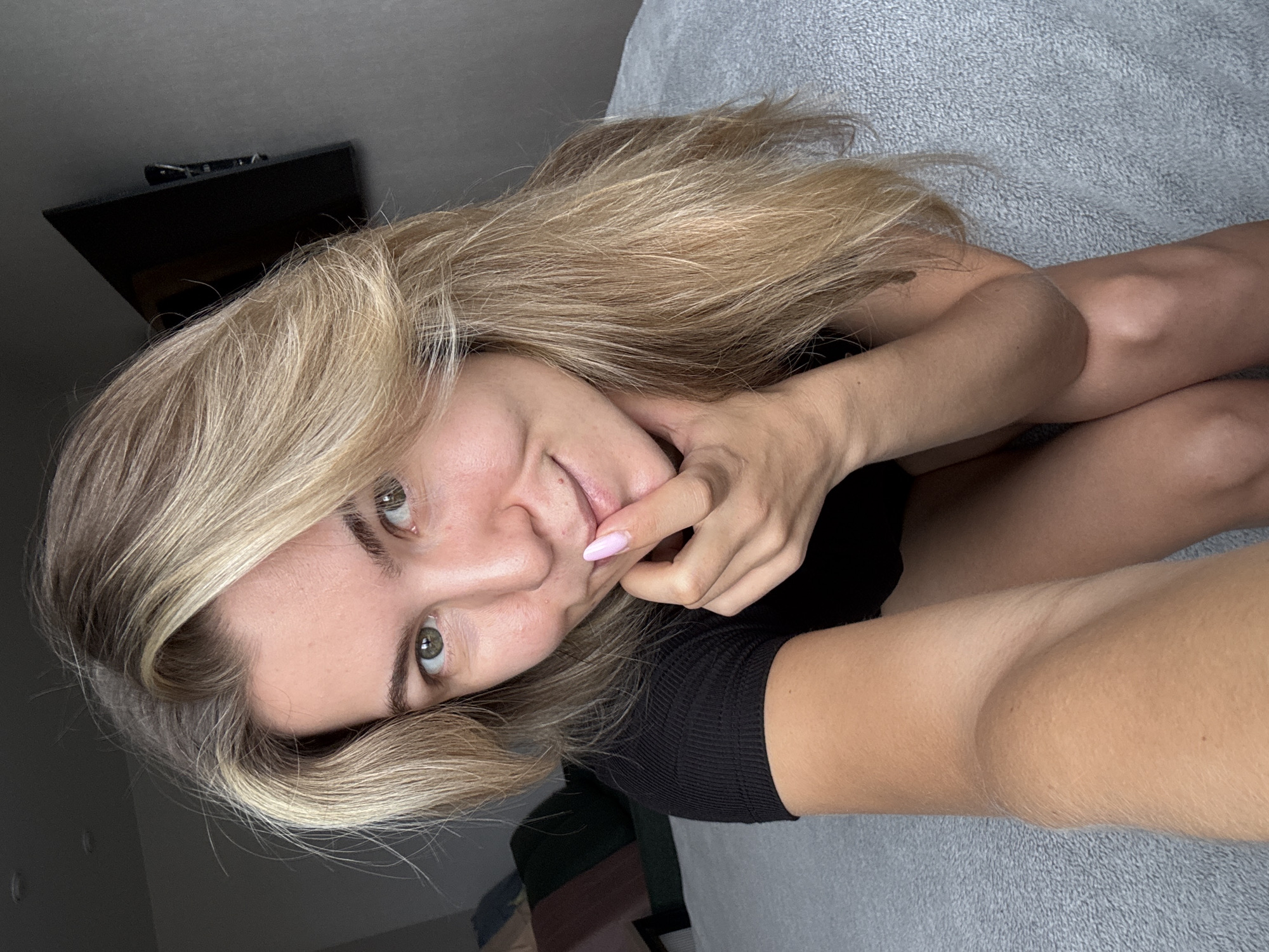 A woman with blonde hair and green eyes sits on a bed, looking at the camera. She is wearing a black top and has her hand to her mouth, with light pink nail polish on her fingernails. The bed is covered with a gray textured blanket. In the background, a dark wall is visible with a black flat-screen television mounted on it. To the left of the woman, a blurry sofa with colorful cushions can be seen. The lighting suggests it is daytime, possibly indoors with natural light.