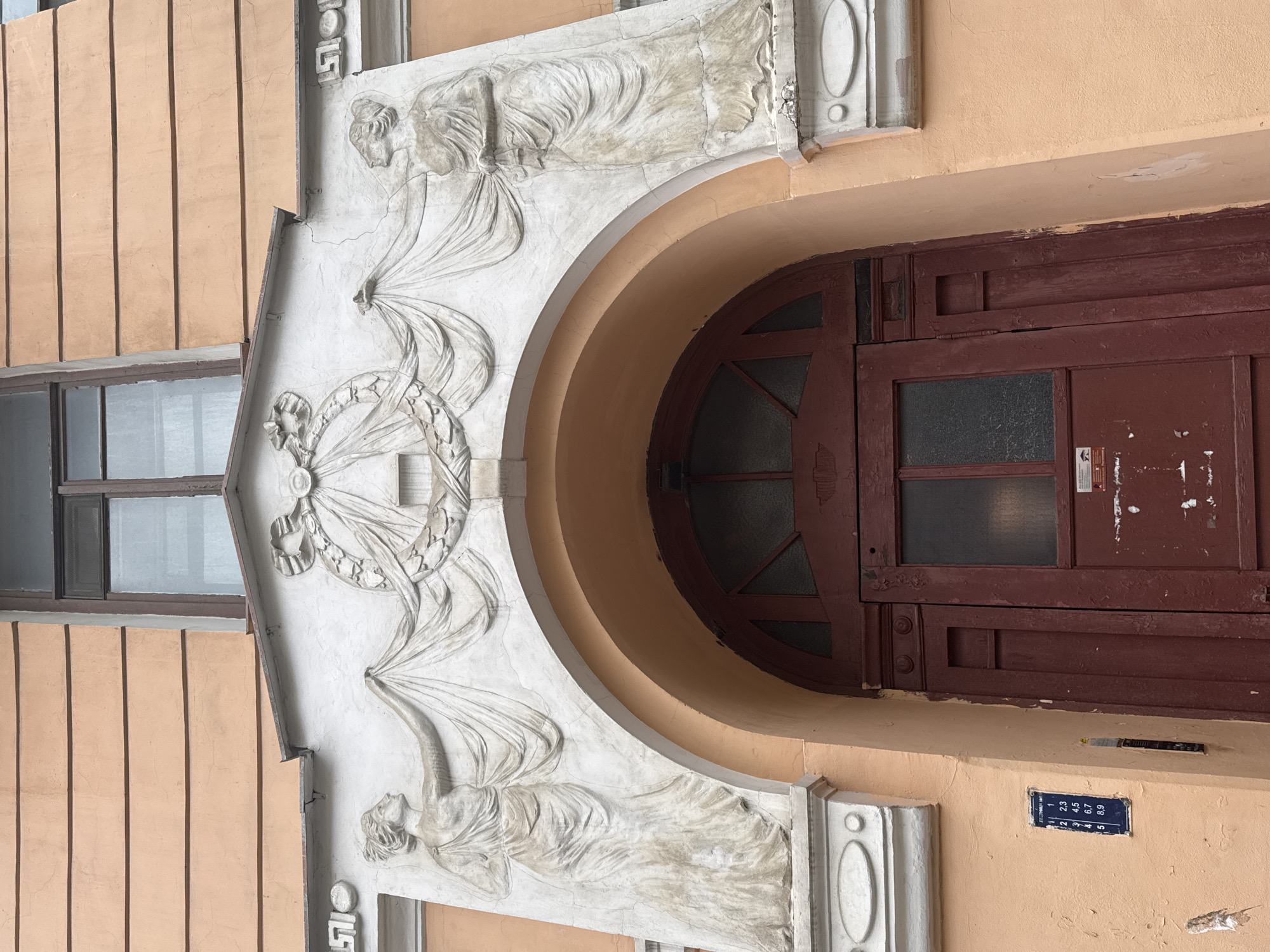 The image shows the entrance to a building in Saint Petersburg, Russia, featuring a classical architectural style.
The main elements are:
* **Entrance:** A round-arched doorway leads into the building. The door itself is made of wood and is dark red or brown. It is weathered, showing signs of peeling paint and disrepair. The door includes a glass panel.
* **Architectural Details:** The entrance is flanked by decorative elements. Above the arch is a keystone-like structure with a wreath or circular design. There are two sculpted figures of women, one on each side of the arch. They are carved in a classical style. The wall is covered with a light-colored stucco finish.
* **Street Number:** A small blue sign with white numbers is visible on the right side of the entrance. It lists the numbers 1-5 and 23,45,67,89.
* **Environment:** The surrounding walls are light orange. A window is seen to the left of the entrance.
The architectural style suggests that the building dates from the late 19th or early 20th century. The scene is in Saint Petersburg, Russia.