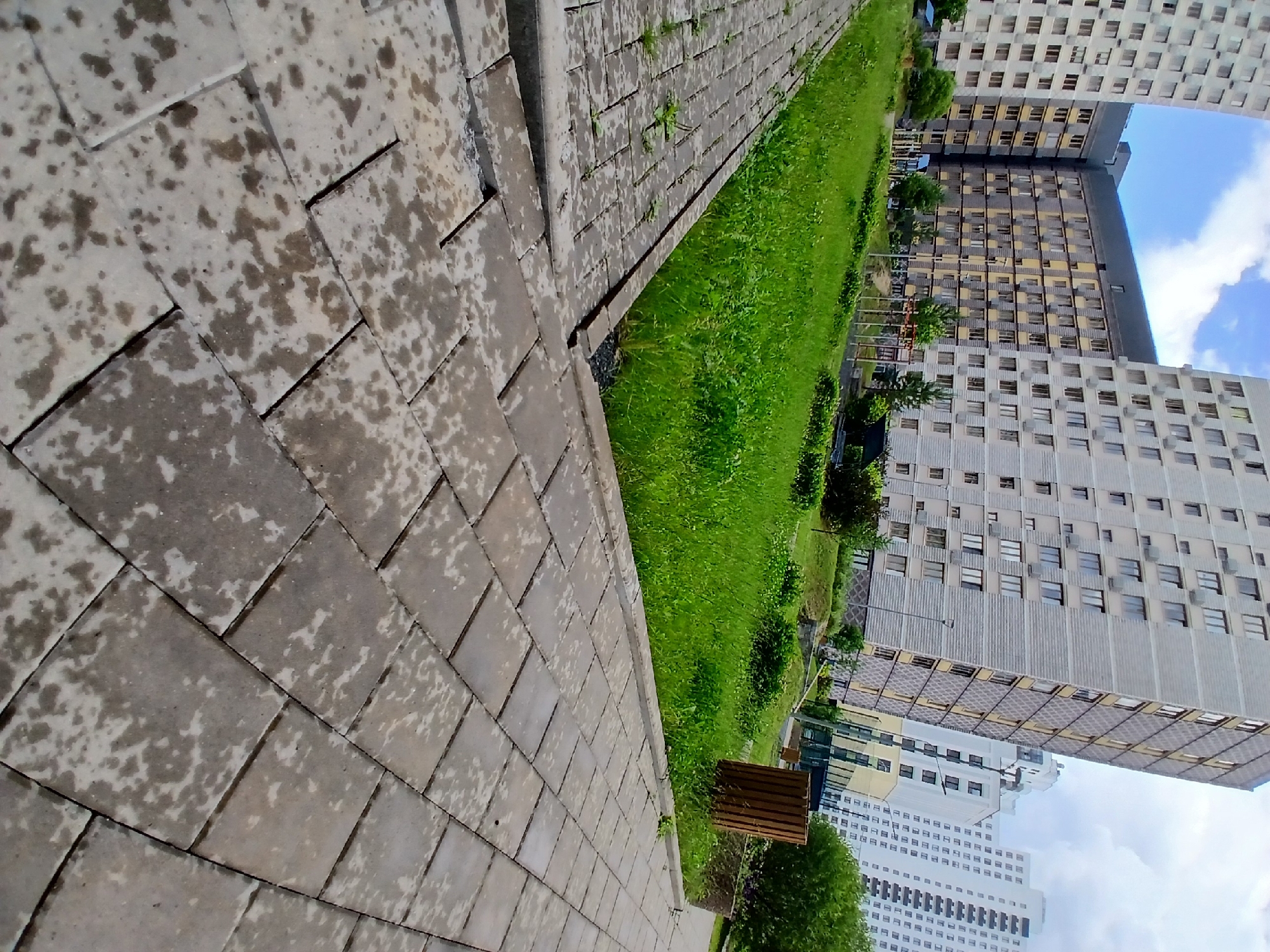 The image depicts a modern residential complex in Yekaterinburg, Russia. The foreground shows a paved walkway made of large, light-colored square tiles, some of which appear wet and discolored, suggesting recent rain. The walkway leads towards a well-maintained green lawn with lush grass, bordered by a concrete curb. Behind the lawn, a series of tall, contemporary apartment buildings are visible. These buildings are characterized by their modular design, featuring multiple windows and balconies. Some buildings are light gray with dark accents, while others are white. The sky above is partly cloudy, with patches of blue. In the middle ground, there are some trees and shrubs, and what appears to be a children's playground with colorful equipment. A wooden trash receptacle is also present near the edge of the lawn. The overall scene conveys a sense of urban development and residential living.