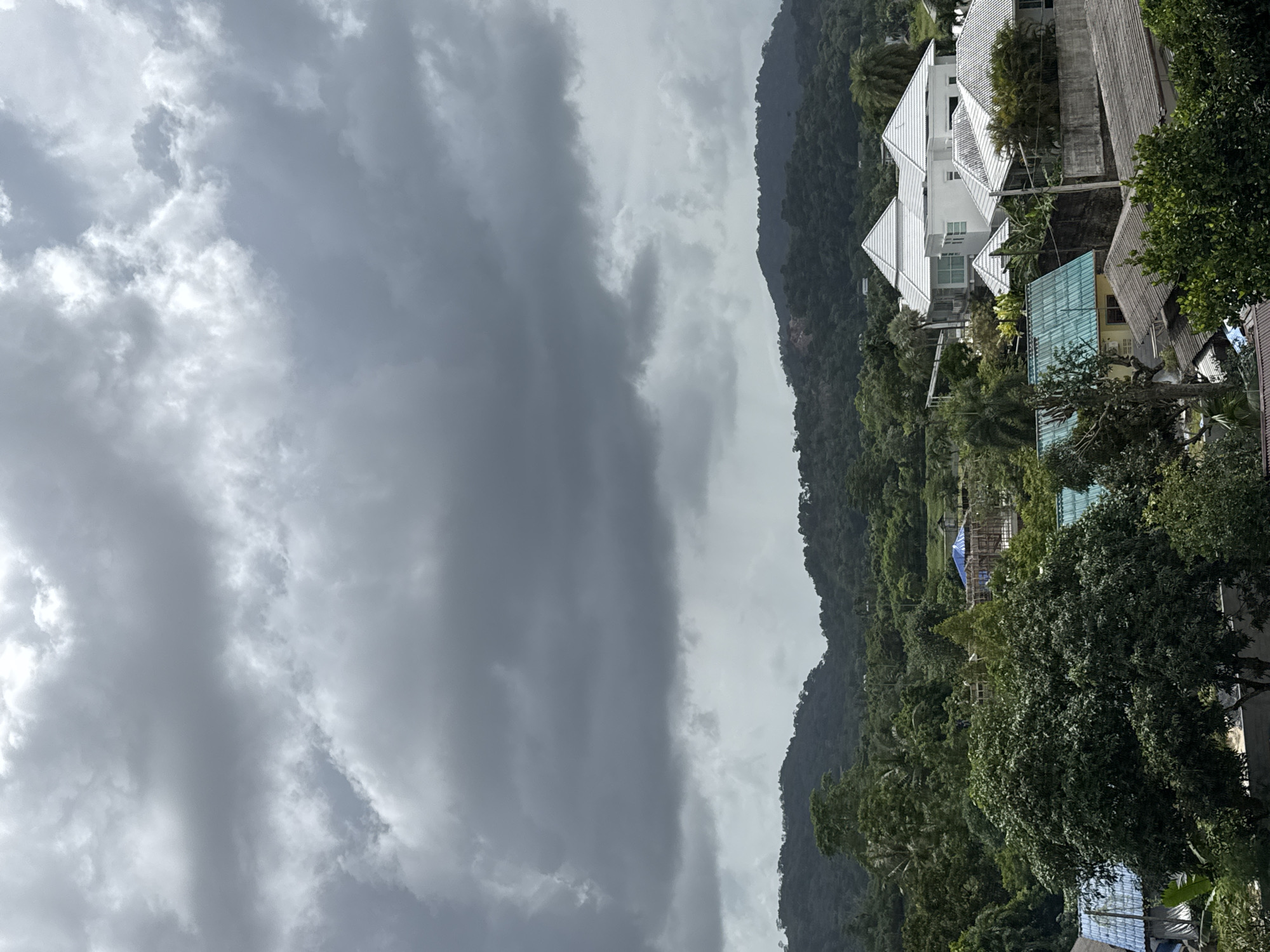 This image, taken from an elevated viewpoint in Kathu, Thailand, captures a cloudy sky over a lush, tropical landscape. The sky is dominated by dark, heavy clouds, suggesting an impending storm. Below the clouds, rolling green hills form the backdrop, densely covered with trees. In the foreground, a residential area is visible with several houses. The houses have distinctive white, angular roofs, and some feature turquoise or gray tiled roofs. Various types of trees, including large, mature ones with dense foliage, are interspersed among the buildings. The overall scene depicts a natural environment with human habitation.