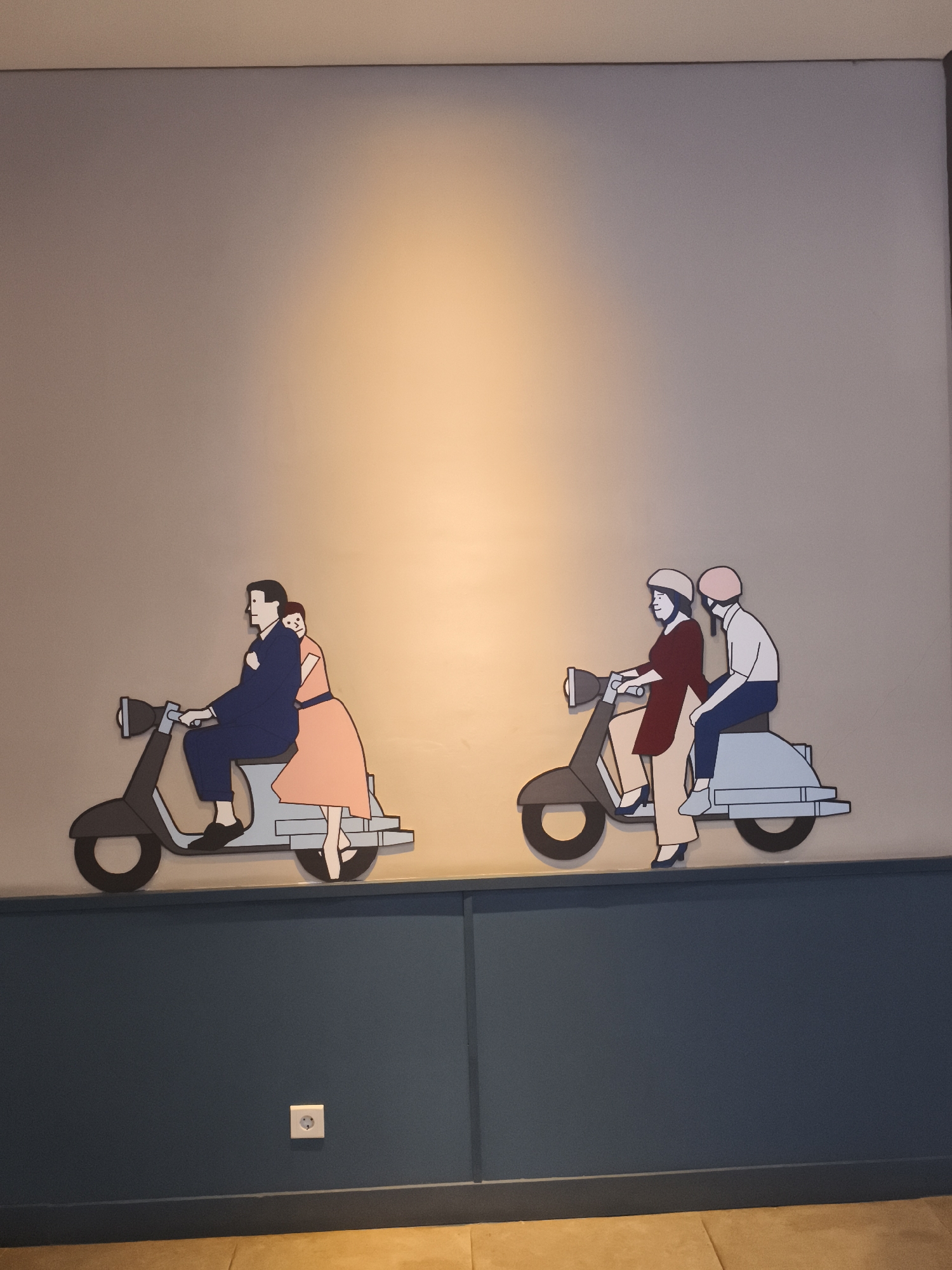 The image displays a wall decorated with two cartoon-style cutouts of people riding scooters. On the left, a man in a blue suit and a woman in a pink dress are depicted on a scooter. The woman is seated behind the man, with her arms wrapped around his waist. On the right, two individuals wearing helmets, one in a maroon top and cream pants, and the other in a white shirt and blue pants, are on another scooter. The wall behind the cutouts is a light, neutral color, with a warm light source illuminating the upper portion. Below the wall art, a dark blue wainscoting is visible, and a white electrical outlet is mounted on it. The scene appears to be indoors, possibly in a public space or restaurant. The presence of a scooter motif, popular in Southeast Asia, could suggest a thematic decoration for such a venue.