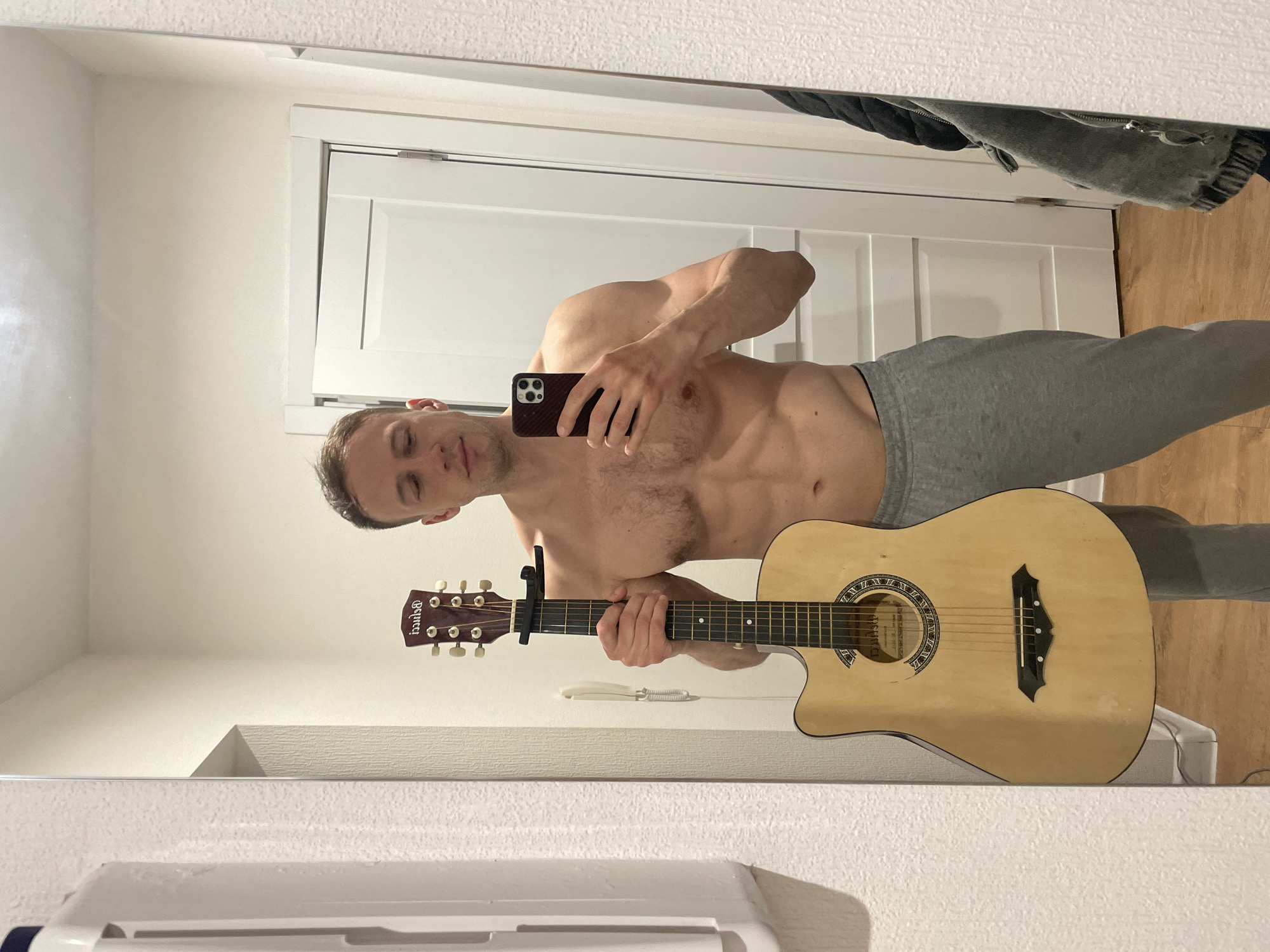 An indoor mirror selfie captures a shirtless man holding a smartphone and an acoustic guitar. The man, who appears to be a young adult with short dark hair and a muscular physique, is wearing grey sweatpants. His right hand holds the dark-cased smartphone to take the picture, while his left hand is positioned on the neck of the guitar. He looks downwards, seemingly at the phone screen, with a neutral expression.
The guitar is a light-colored acoustic model with a cutaway body, and a black capo is clamped to its neck. The brand "Stagg" is visible on the guitar's headstock, which features six tuning pegs.
The background, visible through the mirror, includes a white paneled door, light-colored walls, and a textured white ceiling or upper wall section. A dark piece of clothing, possibly a jacket or hoodie, is visible hanging in the upper right corner. The floor reflected appears to be light-colored wood. The lighting is artificial indoor light, casting soft shadows. The scene suggests a casual setting, possibly a bedroom or living area, in Saint Petersburg, Russia.