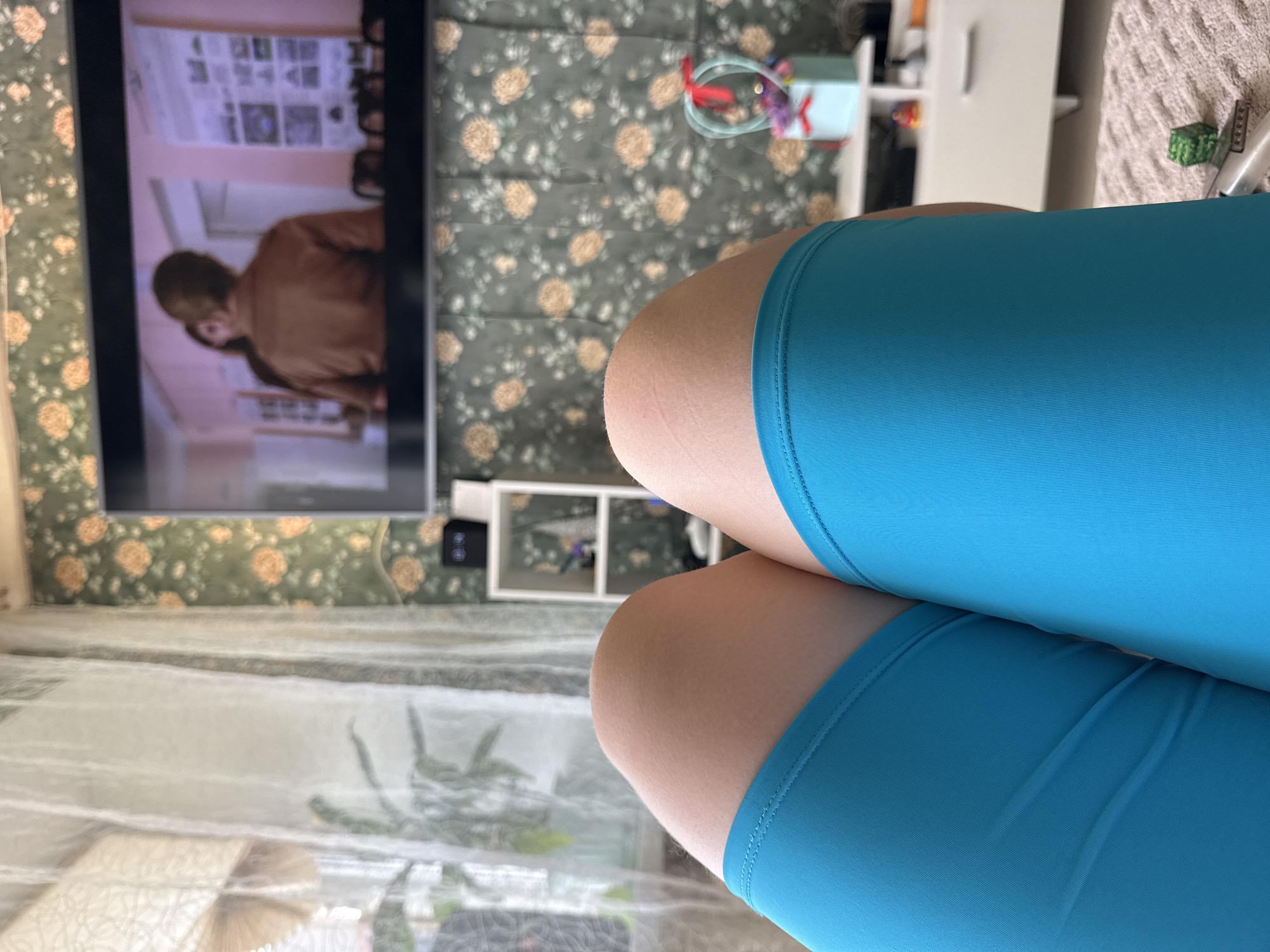 The image depicts a first-person perspective of someone sitting in a living room, wearing bright blue shorts. The focus is on the person's legs in the foreground. In the background, a television screen displays a scene from a movie or show, featuring two individuals with their backs to the camera. The room's decor includes floral wallpaper and a white shelf unit with decorative items. A window with sheer curtains is visible on the left side. The scene is set indoors, likely in a residential apartment in the city of Kopeysk, Russia, given the user's context.