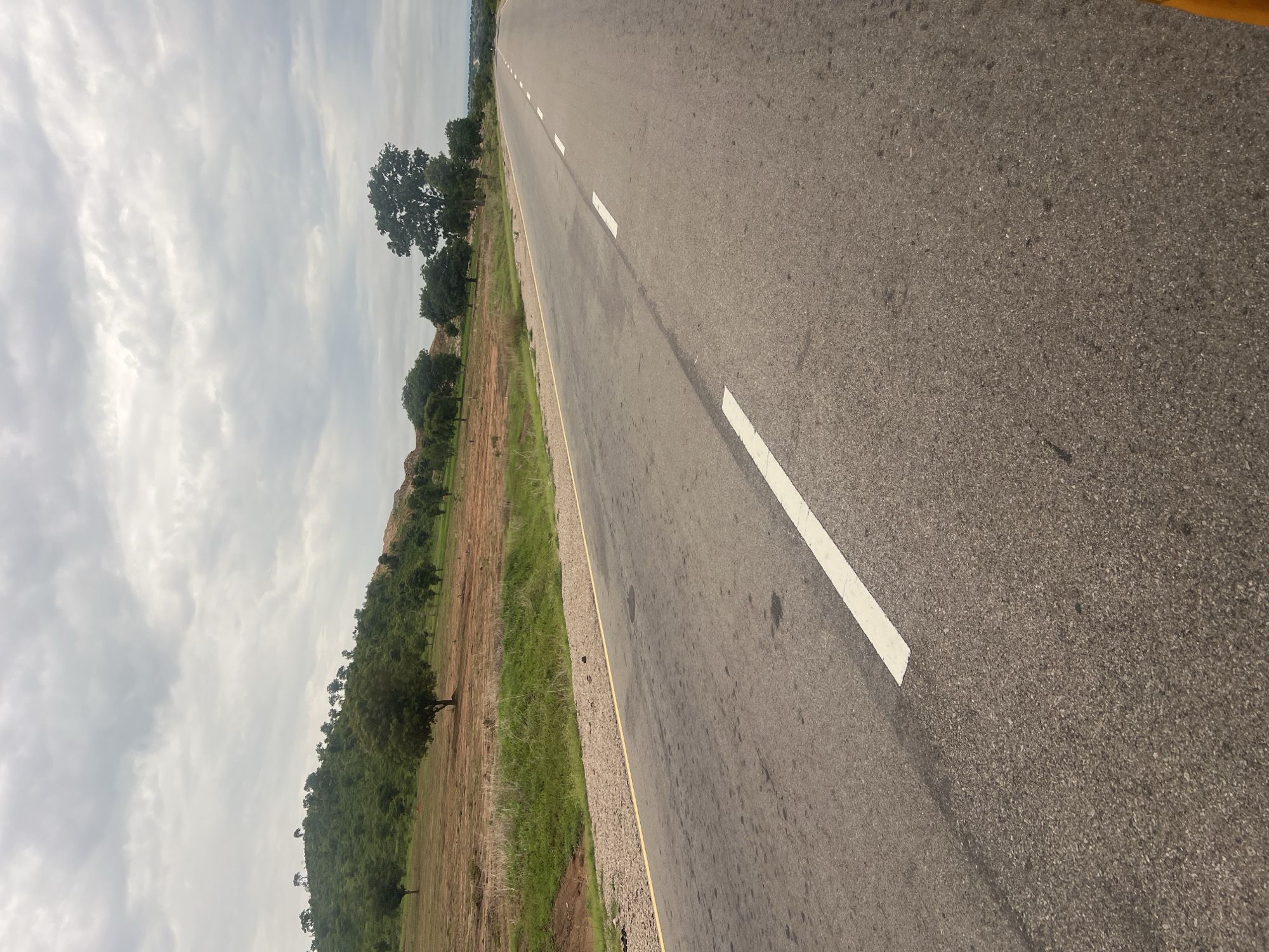 This is a vertical, eye-level, outdoor shot of an asphalt road in Minda, Nigeria. The road stretches into the distance, marked by intermittent white lines on its right side. To the left of the road, there's a grassy verge, followed by dry, brown earth and a lush green hillside dotted with trees. The sky is overcast with soft, wispy clouds. The overall impression is one of a quiet, rural landscape under a subdued sky. There are no people or animals visible in the frame. The lighting suggests it is daytime, likely mid-morning or afternoon, given the lack of strong shadows. The scene evokes a sense of journey and the open road, with a touch of natural beauty in the surrounding landscape. No text is visible in the image.