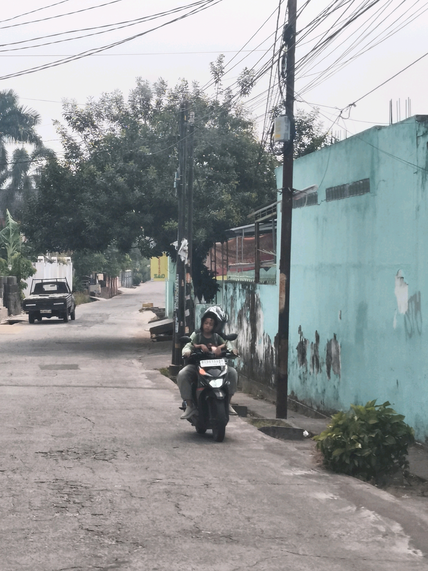 The image depicts a residential street in Palembang, Indonesia, on an overcast day. The road surface is uneven, with visible cracks and patches of wear. On the right side of the street, a light blue building with peeling paint and graffiti stands adjacent to a wooden utility pole adorned with numerous electrical wires. In front of the building, a small, dense green bush grows.
A motorcycle with two occupants is traveling away from the camera, positioned on the right side of the road. The rider, wearing a helmet, is visible in the foreground, and the passenger, also wearing a helmet, is seated behind. The motorcycle's license plate reads "BD3813ARP".
Further down the street, on the left, a black pickup truck is parked. Beyond the truck, there are more buildings and trees, suggesting a typical urban or suburban neighborhood. The overall atmosphere of the image is mundane, capturing a slice of everyday life in Palembang.