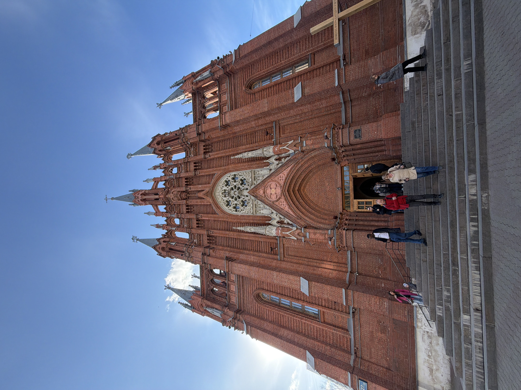 A low-angle, outdoor shot of a red brick cathedral, likely in Moscow, Russia, given its architectural style. The building's facade features a large rose window, pointed arches, and several spires reaching towards a clear blue sky. People are standing on the stone steps leading to the entrance. The steps are a gray, contrasting with the red brick. The sun is positioned behind the building, creating a slight glare. A wooden cross is seen near the top right of the building.
