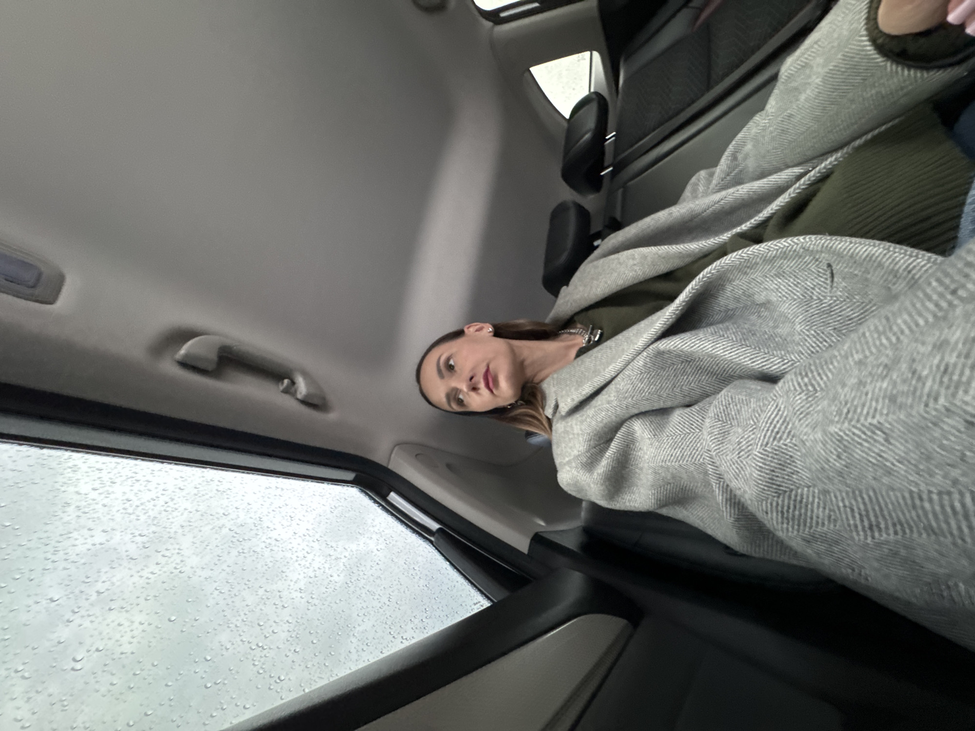 The image appears to be taken inside a car, possibly in Sochi, Russia, due to the style of vehicle common there. Rain is visible on the car window. A woman is in the passenger seat, wearing a grey herringbone patterned coat. The interior of the car is grey. The seat behind the passenger appears to be black leather. The photo is taken from an unusual angle looking upwards.