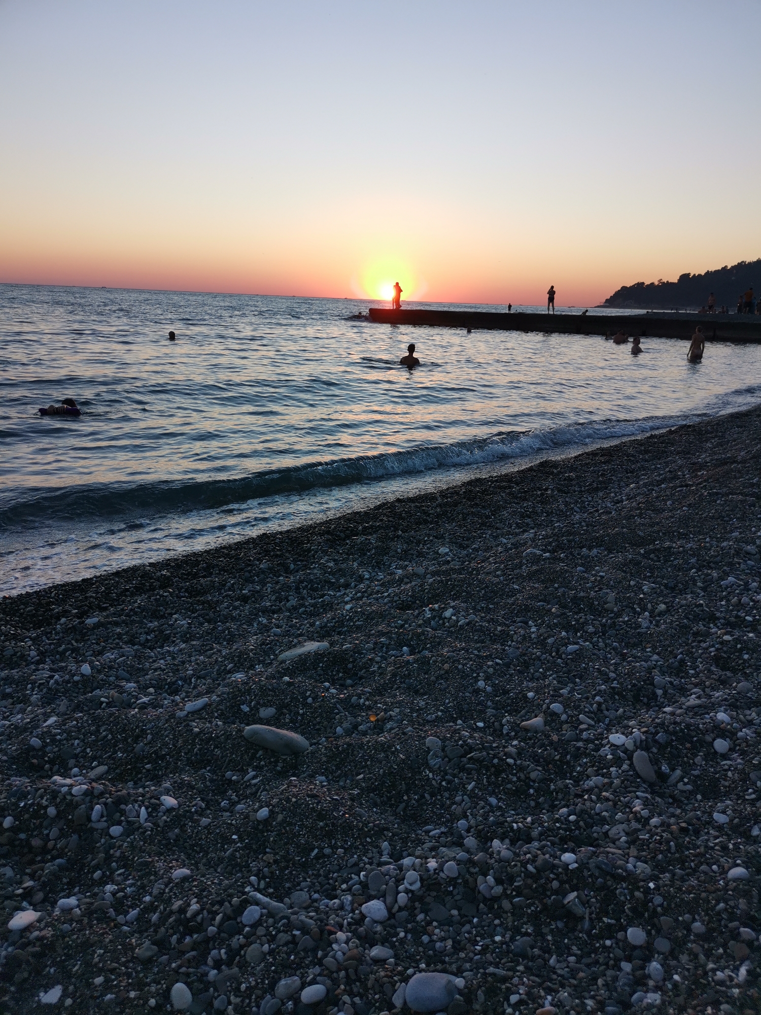 The image depicts a serene sunset scene at a pebble beach. In the foreground, a dark-colored pebble beach stretches towards the water. The sea is calm, with gentle waves lapping at the shore. Several individuals are visible in the water, some swimming and others standing. In the mid-ground, a pier extends into the sea, with a few silhouetted figures standing on it. The sun is setting on the horizon, casting a warm orange glow across the sky and reflecting on the water. A distant, tree-covered hill is visible on the right side of the image. The overall atmosphere is peaceful and idyllic. The location is specified as Bogushevka, Russia.