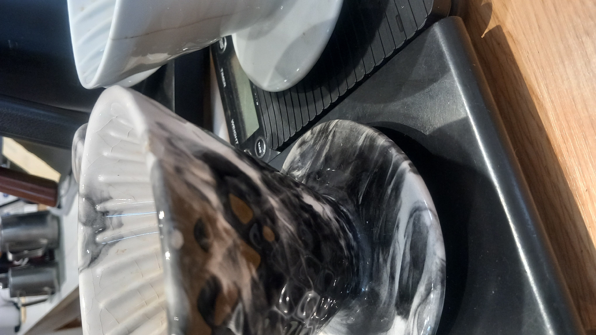 The image shows a close-up of a coffee brewing setup. In the foreground, a marbled black and white ceramic pour-over cone sits on a black digital scale. To the right, a white ceramic pour-over cone rests on its saucer. In the blurred background, metal pitchers and a wooden object are visible, suggesting a kitchen or cafe environment. The image does not provide any information about the city of Kos, Greece.