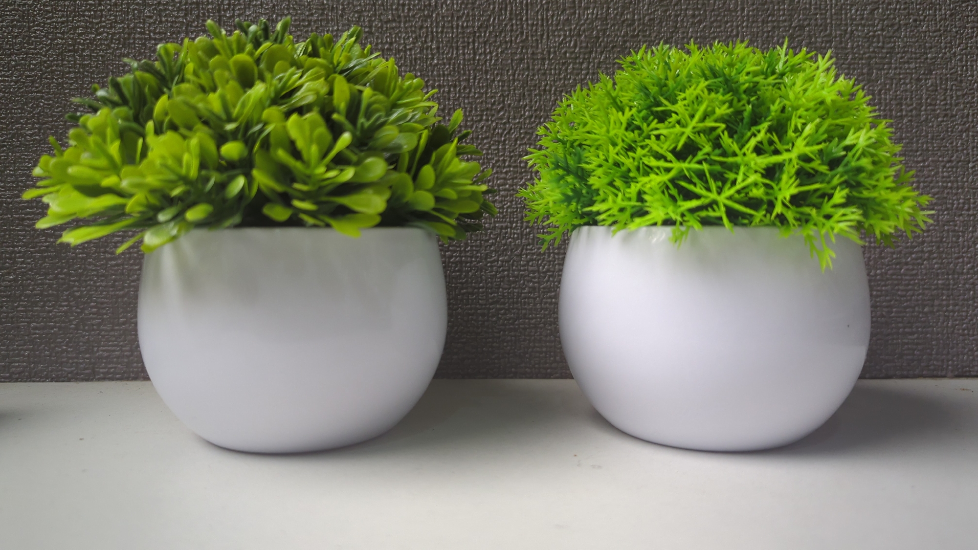 The image shows two artificial potted plants. The plant on the left has dense, rounded green foliage. The plant on the right has finer, feathery green foliage. Both plants are in white, spherical pots. The pots are placed on a white surface, likely a shelf or table. Behind the pots is a textured gray wall. The lighting is neutral, casting soft shadows. The image appears to be taken indoors. There is no visible indication of the location being in Penjor, Indonesia; this information is extraneous to the visual content of the image.