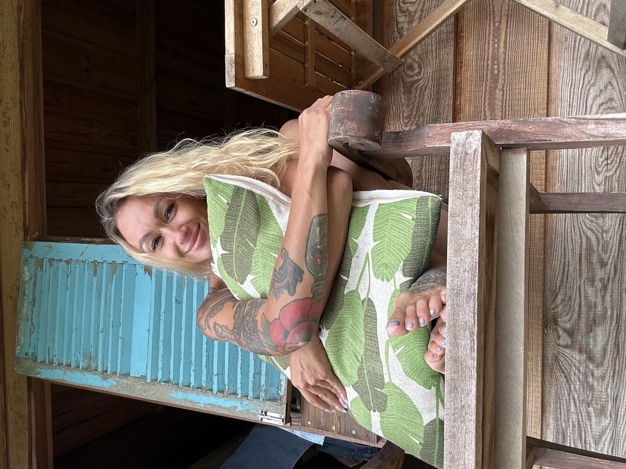 A blonde woman with tattoos on her arms sits in a wooden chair, holding a pillow with a green leaf pattern. She is smiling at the camera. She is wearing a white top and jeans. The background is a wooden wall with blue shutters. The image appears to have been taken outdoors. The location is described as Thon 3, Vietnam.