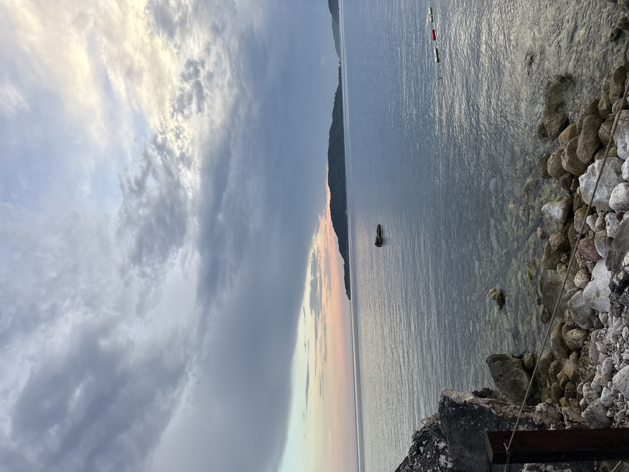 A picturesque twilight scene unfolds at the rocky shore of Kamenovo, Montenegro. The sky above is a canvas of dramatic grey clouds interspersed with streaks of soft pink and yellow, hinting at the setting sun. The calm, serene sea reflects the muted sky, with a few scattered buoys visible on the surface. In the distance, a dark, tree-covered island or peninsula juts out into the water. The foreground is dominated by a rocky beach, with various sizes of stones and pebbles leading down to the water's edge. A rough, rustic wooden post with a rope attached to it stands to the left, adding a touch of human presence to the natural landscape. The overall atmosphere is one of peaceful tranquility.