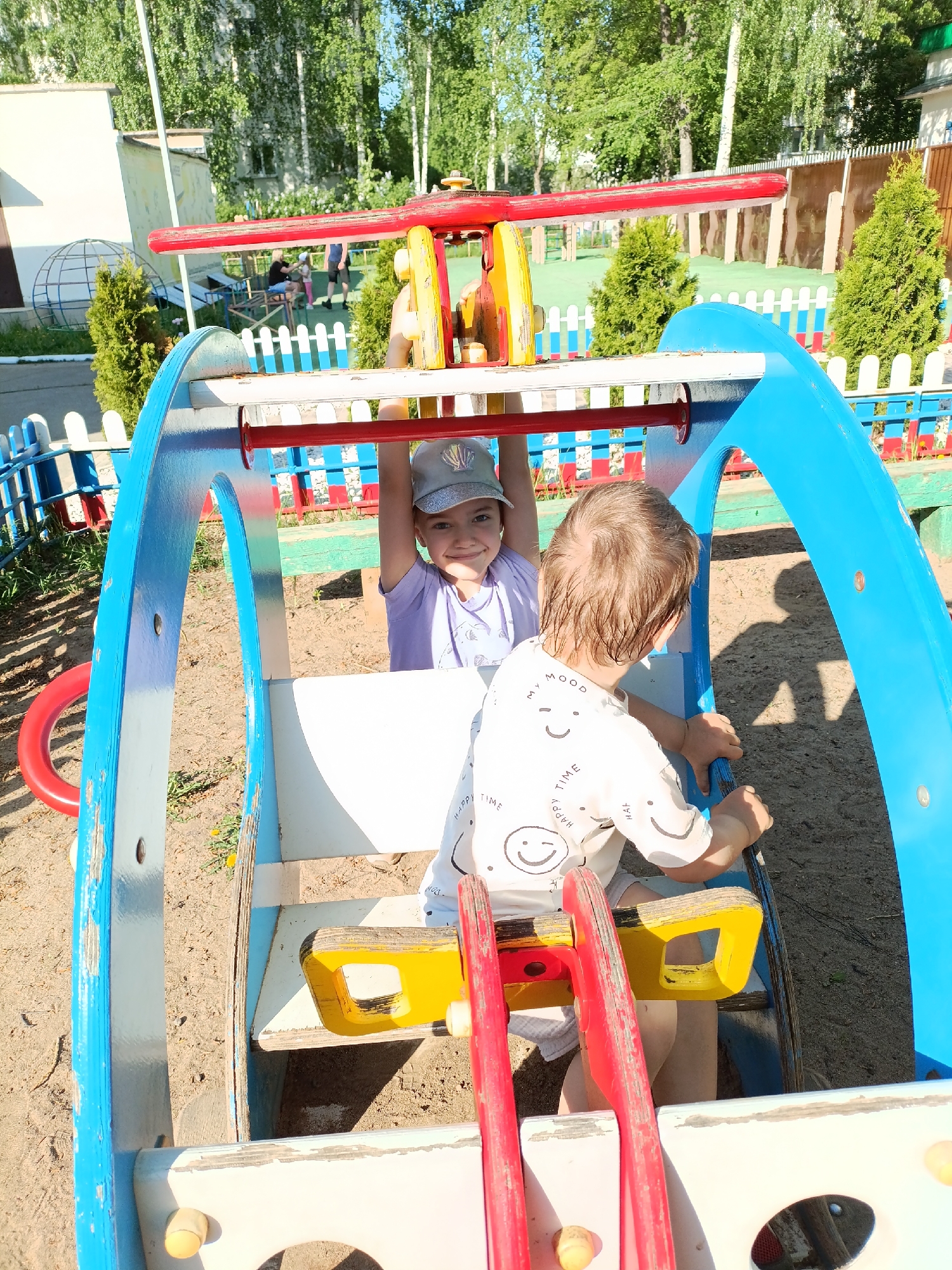 Observed at a playground in Chaikovsky, Russia, are two children interacting with a colorful play structure resembling a helicopter. One child, wearing a light purple shirt and a baseball cap, is positioned in front, holding onto a horizontal bar with an upward gaze and a slight smile. The other child, donned in a light-colored shirt with a smiley-face print, stands behind, appearing to manipulate a part of the structure. The helicopter-shaped structure is multi-colored, featuring shades of blue, red, and yellow. It stands on a sandy ground surrounded by a low fence with a blue and white color scheme. In the background, there are trees and shrubs, suggesting a park-like environment. Another adult and child are visible behind a white picket fence. A building and globe frame can be seen to the left.