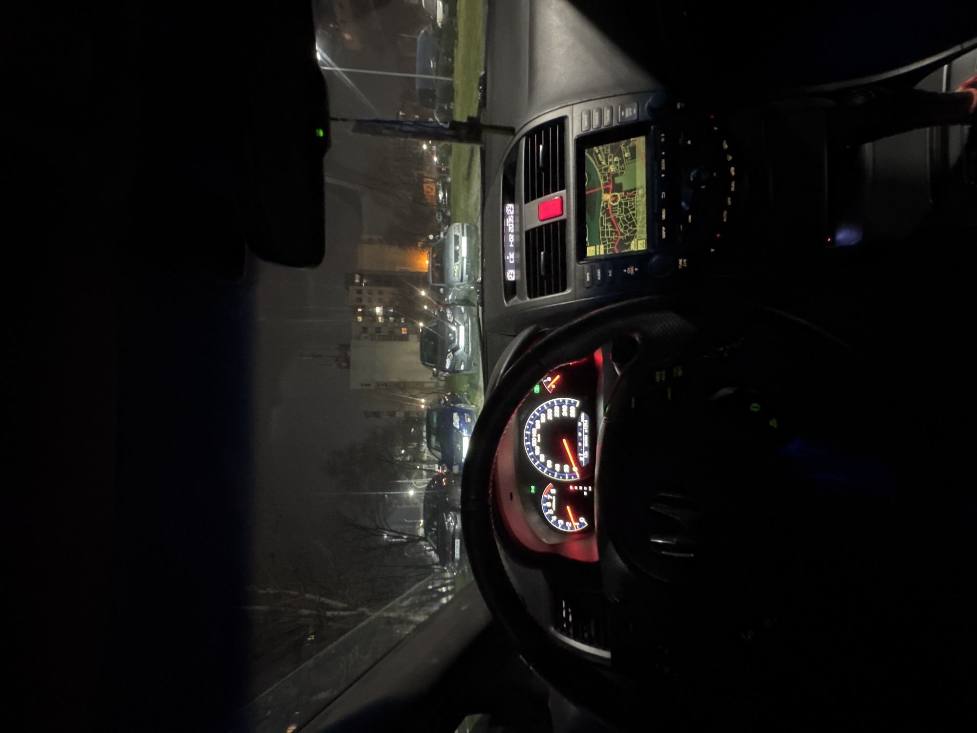 The image is taken from inside a car at night. The dashboard is visible, illuminated with the speedometer showing around 120 km/h and the tachometer indicating a relatively low RPM. A GPS navigation system is displaying a map. Outside the car, there are streetlights, trees, and other parked vehicles visible. The location is likely Plovdiv, Bulgaria, based on contextual information, though specific visual markers for definitive location within the city are not clearly discernible.