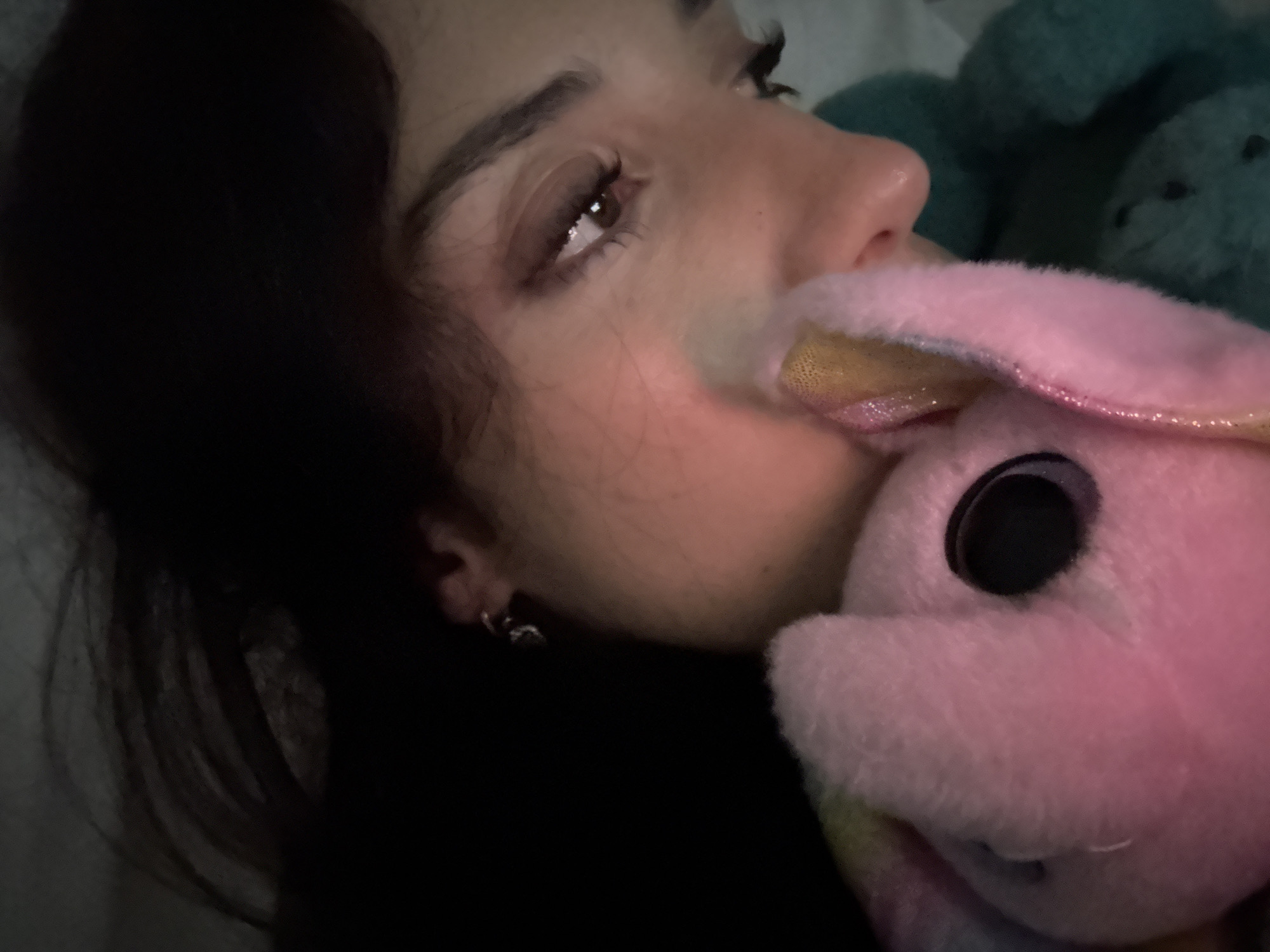 A close-up indoor shot shows a woman lying down, facing slightly upwards and to the right. Her dark hair is visible to the left, and her face is in soft focus. She has dark eyebrows and eyelashes. The woman is holding a pink plush rabbit toy close to her face, partially obscuring her mouth. Behind the pink rabbit, blurry blue stuffed toys are partially visible. The lighting is dim and appears artificial. The location in Buenos Aires, Argentina is impossible to determine from the visual context provided.
